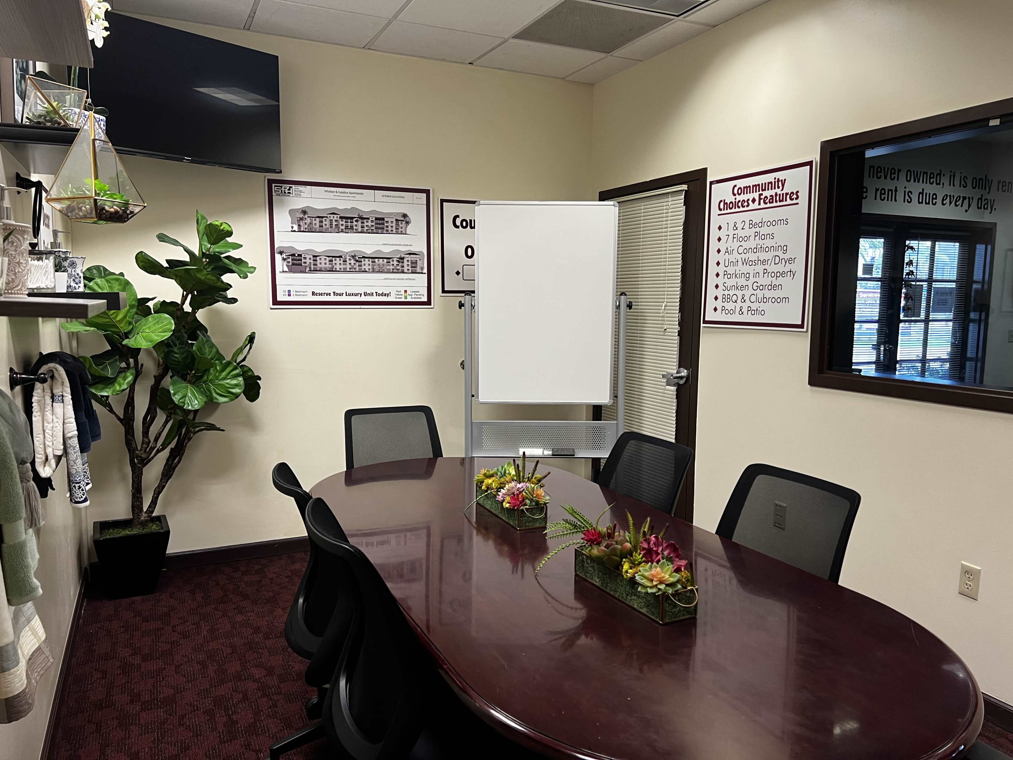 A conference room with a large oval table, several chairs, a whiteboard, and decorative plants on the walls.