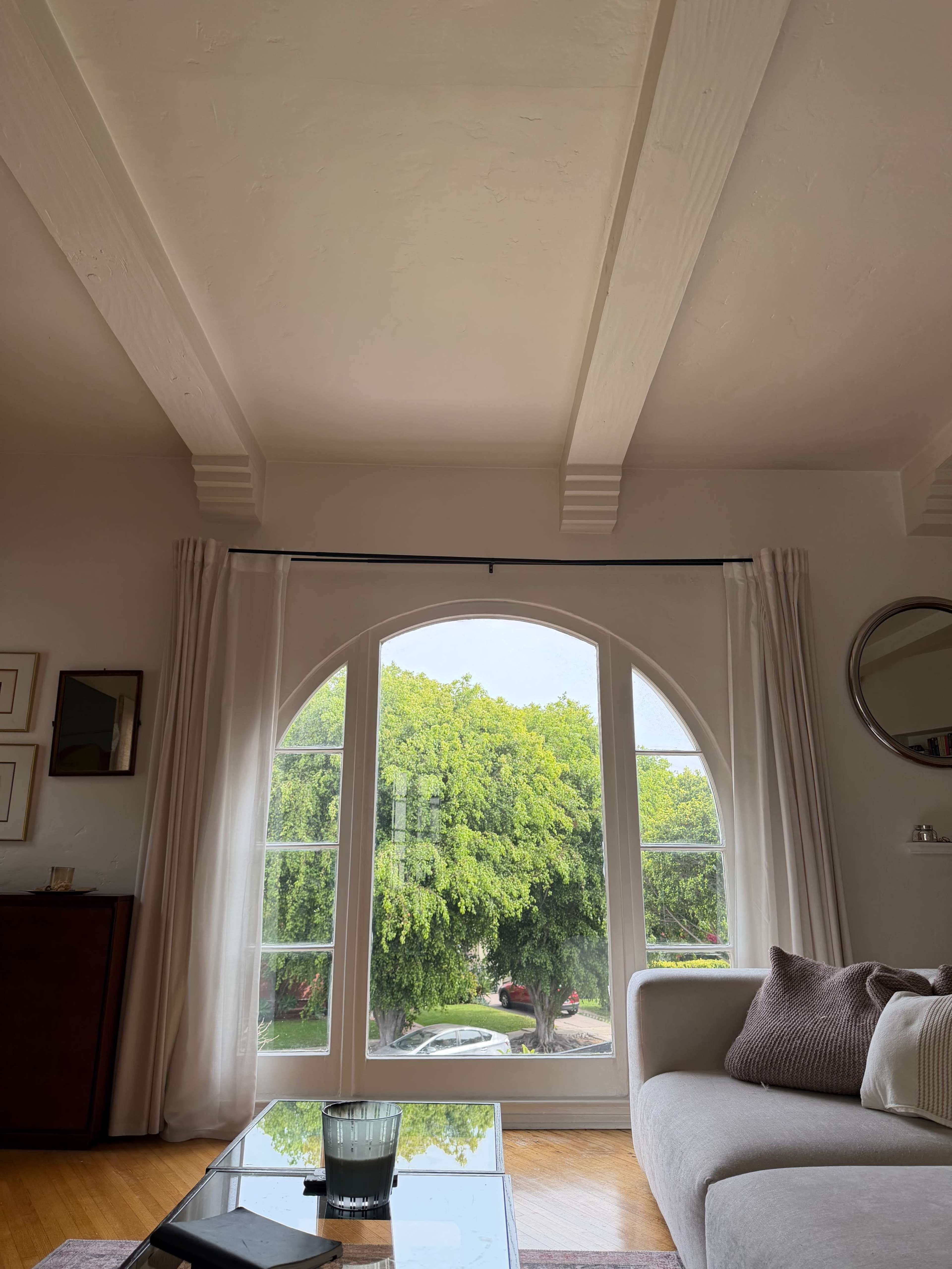 The image shows a room with a large arched window framed by light-colored curtains, revealing green trees outside.