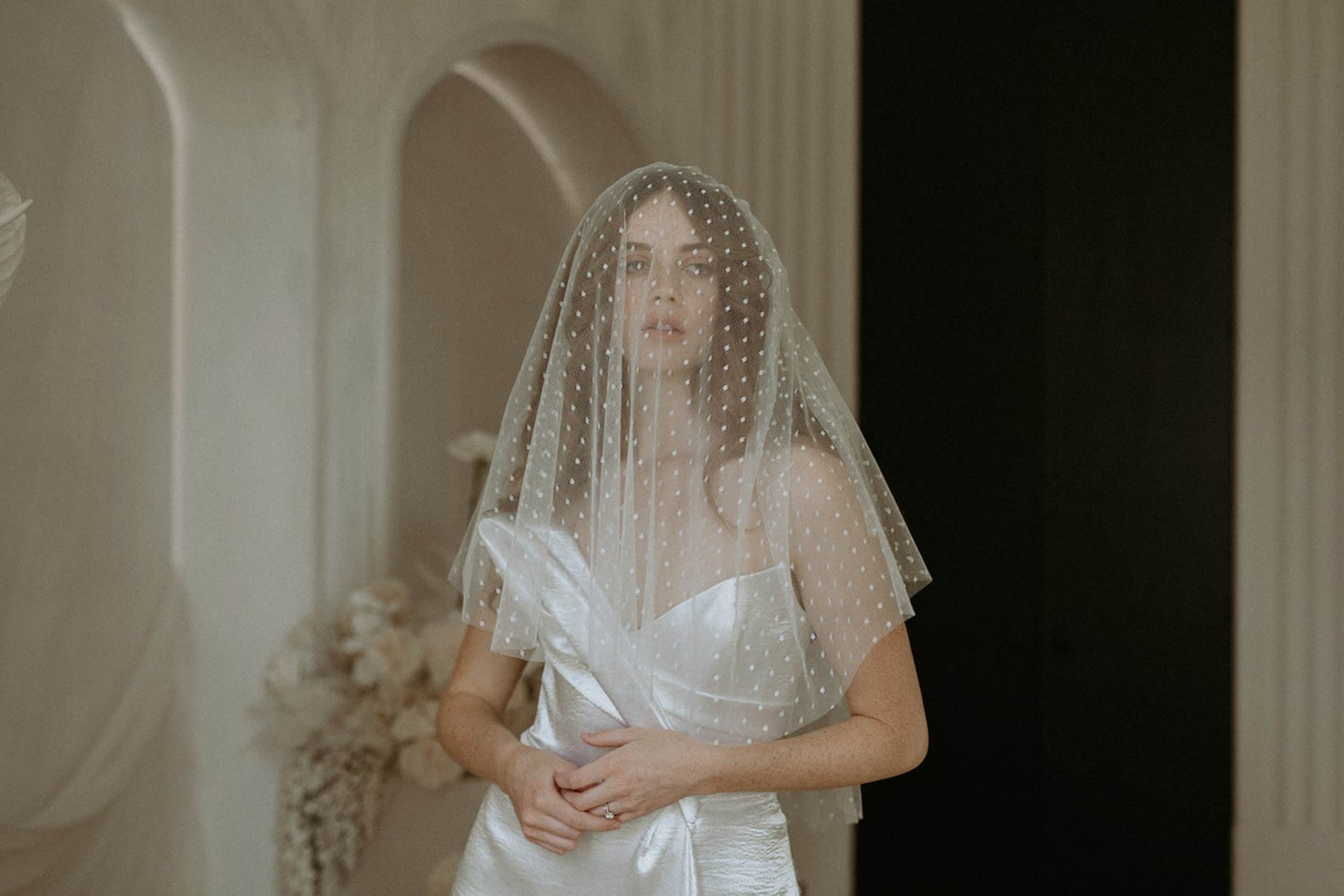 A bride wearing a polka-dot veil stands elegantly in a softly lit indoor setting.