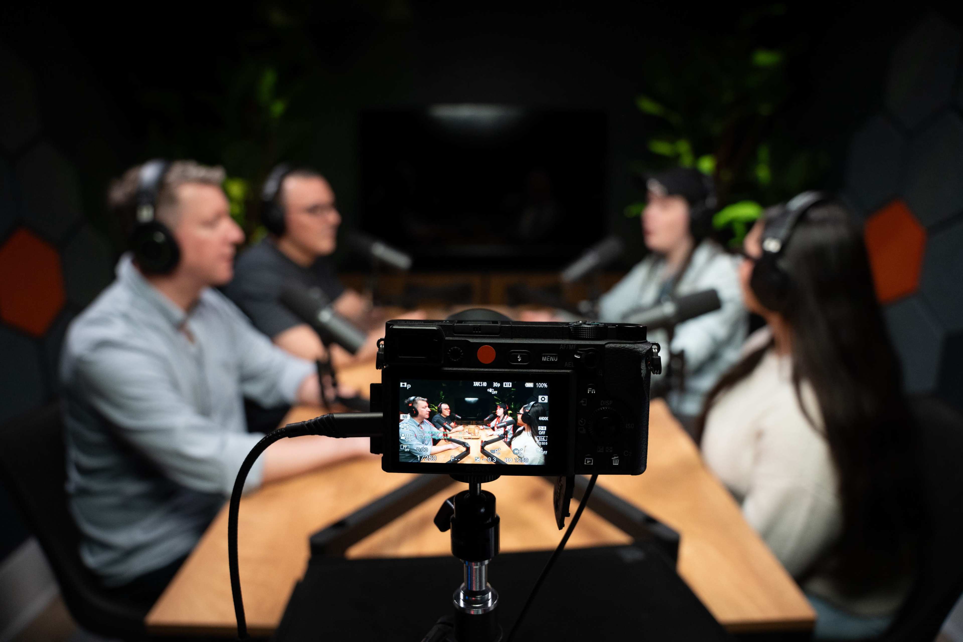 A group of four people are seated around a table, wearing headphones and discussing, while a camera on a tripod captures the scene.