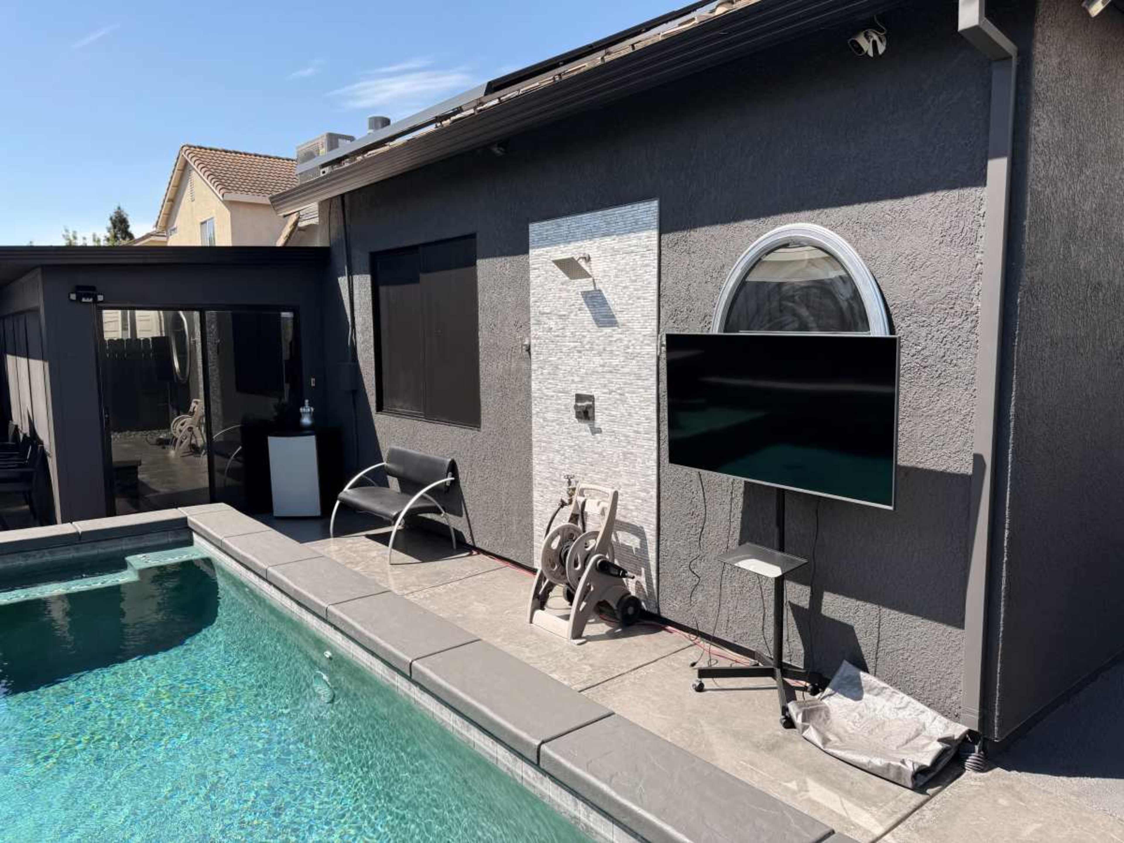 The image shows a pool area featuring a television mounted on a stand next to a gray house, with patio chairs and a clear blue pool in the foreground.