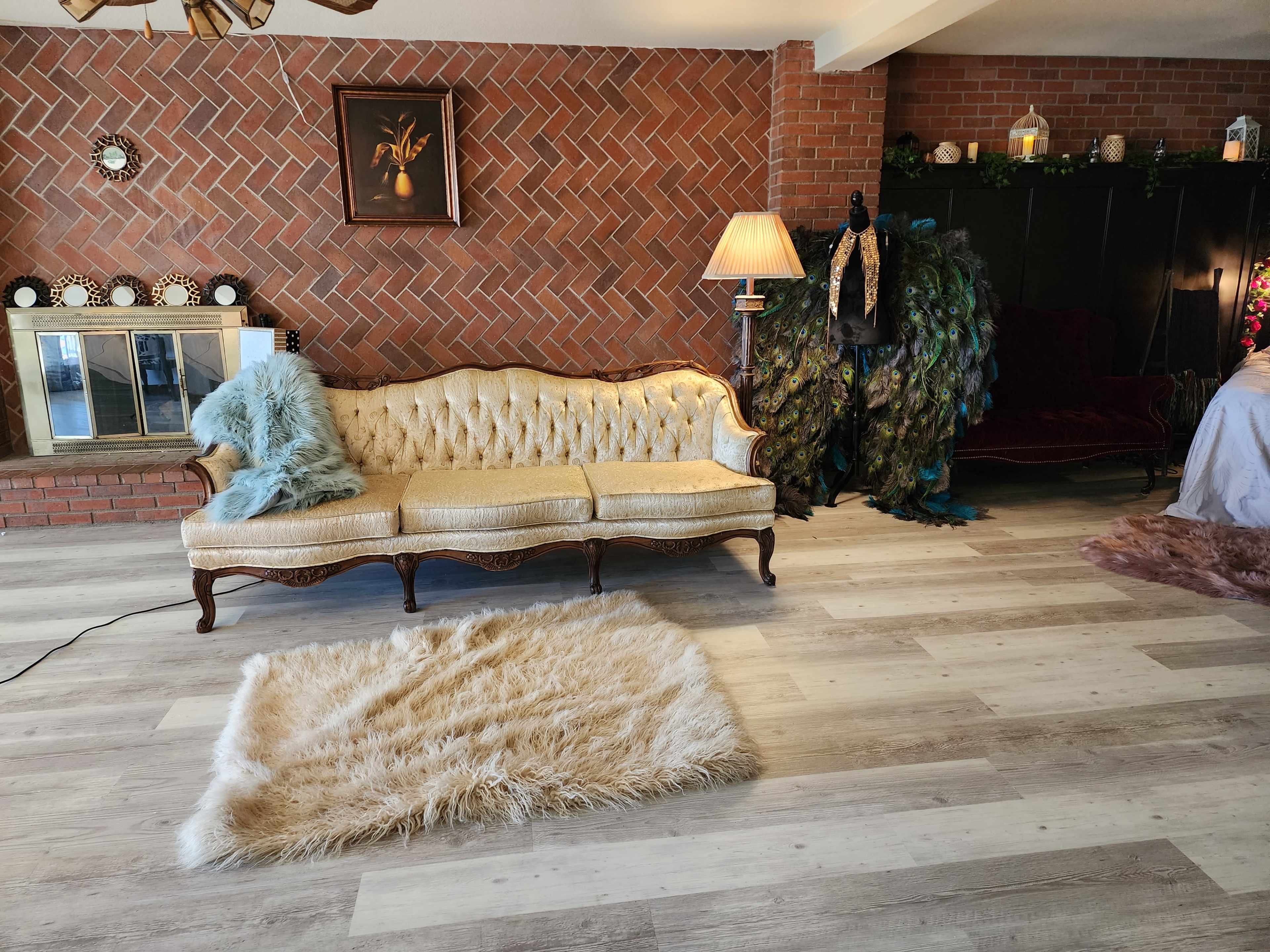 The image shows a vintage gold-colored couch with a light blue throw pillow, placed on a light-colored wooden floor, alongside a fluffy rug and a brick wall backdrop with various decorative items.