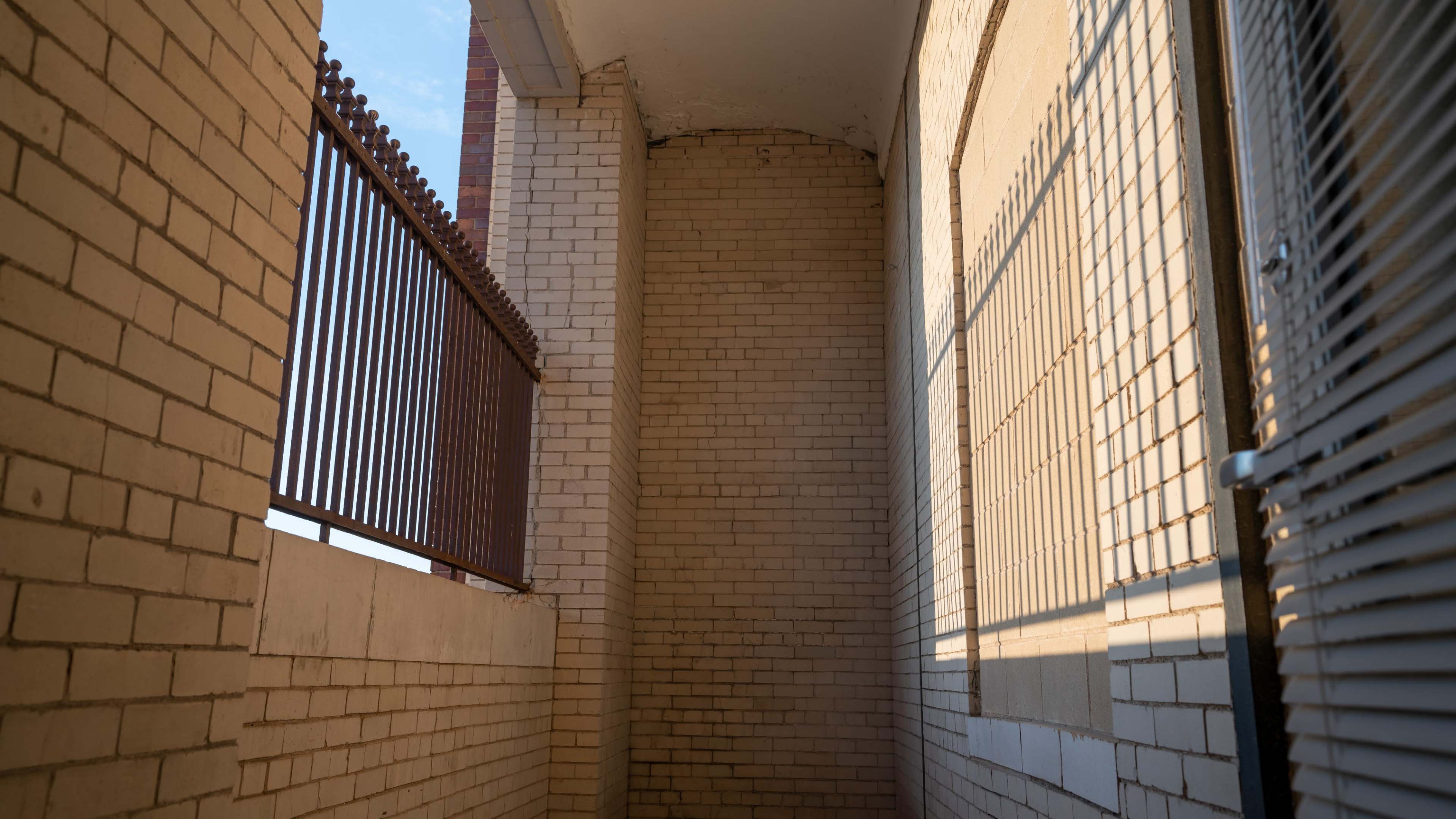 A narrow corridor with brick walls, a metal railing above, and shadows cast by the railing on the wall.