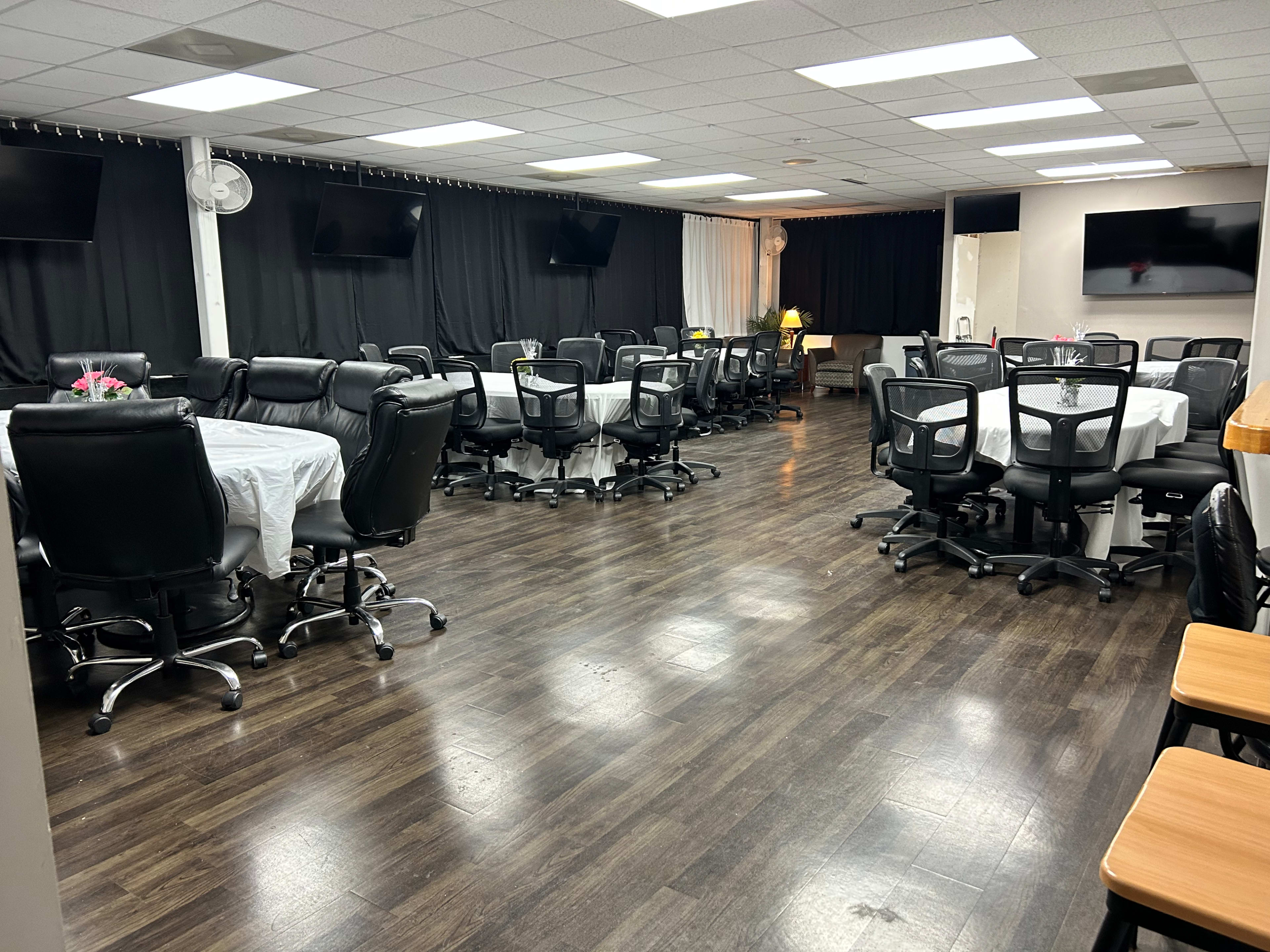 A spacious meeting room with several tables covered in white tablecloths and black rolling chairs arranged around them.