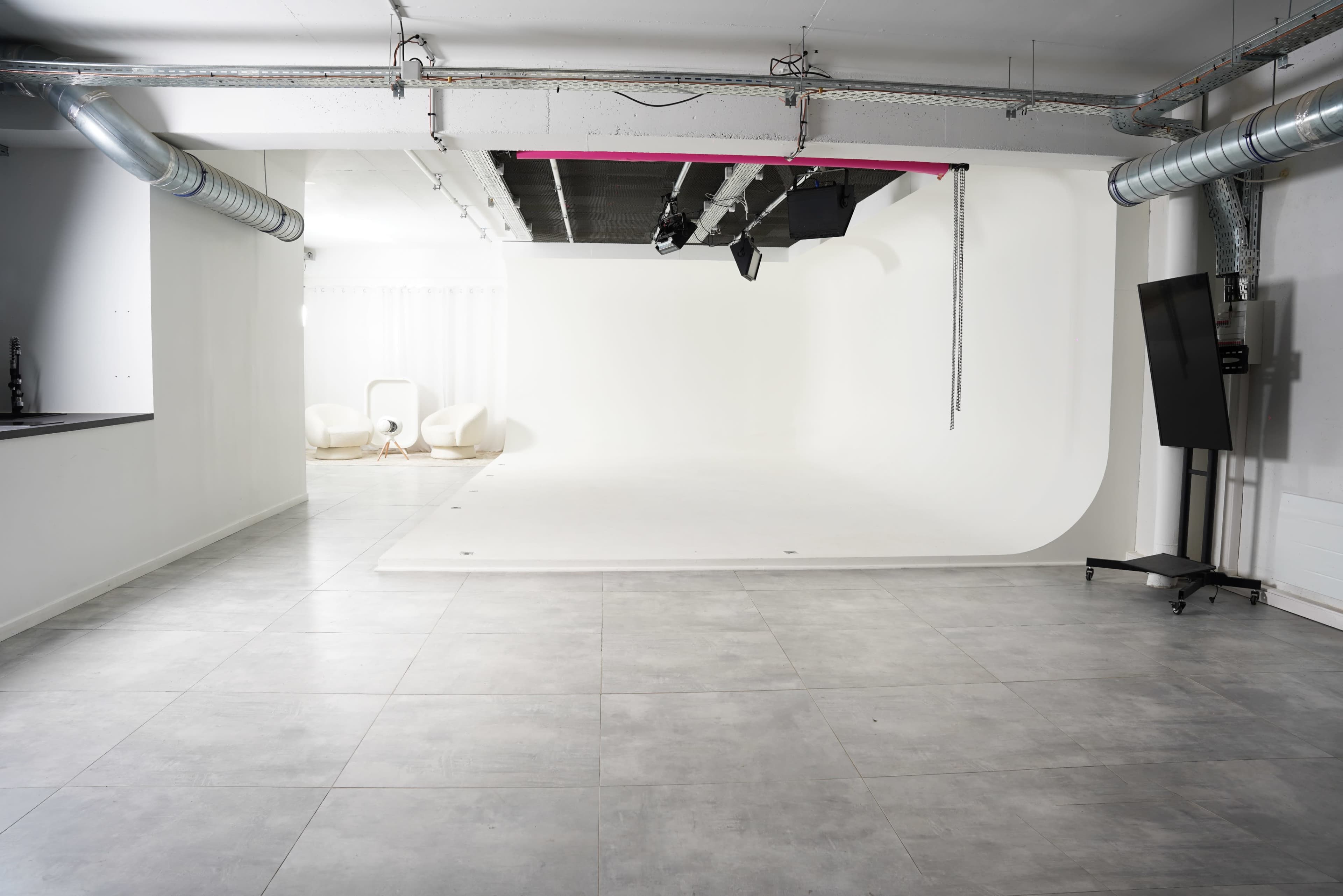 A spacious photography studio with a seamless white backdrop and a light setup, featuring grey tiled flooring and modern furniture in the corner.