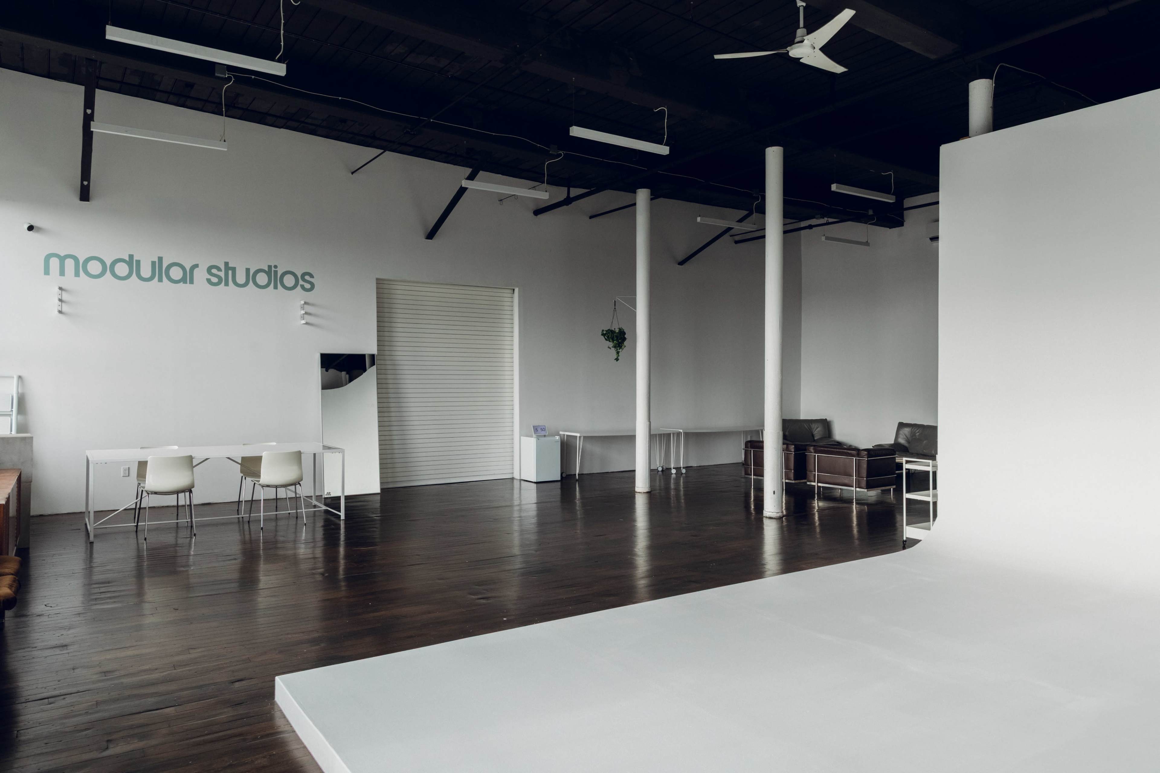 The image shows a spacious studio interior with a minimalist design, featuring white walls, wooden flooring, and a large white backdrop for photography or video.