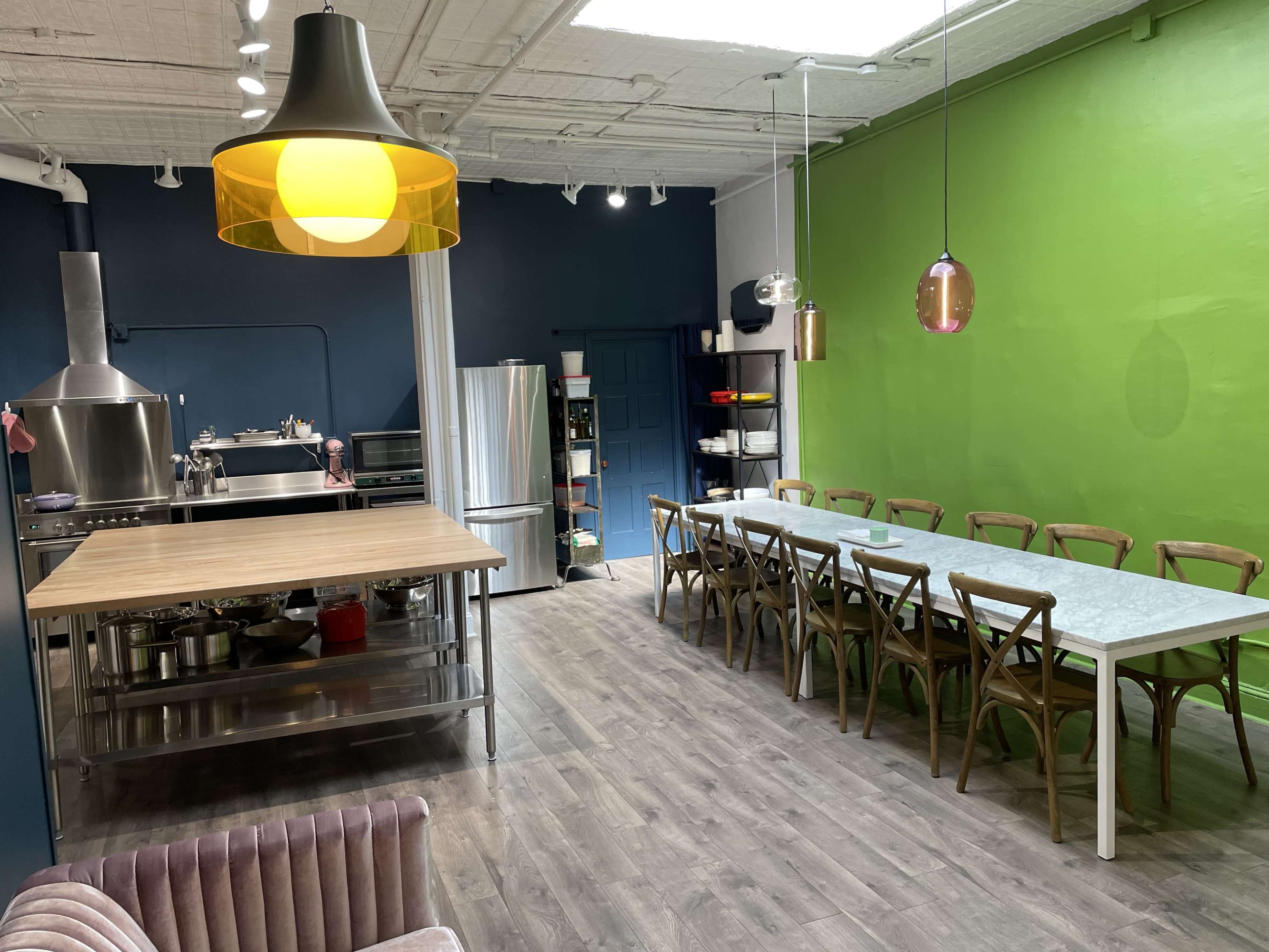 A modern kitchen and dining area features a large table surrounded by wooden chairs, stainless steel appliances, and a vibrant green wall.