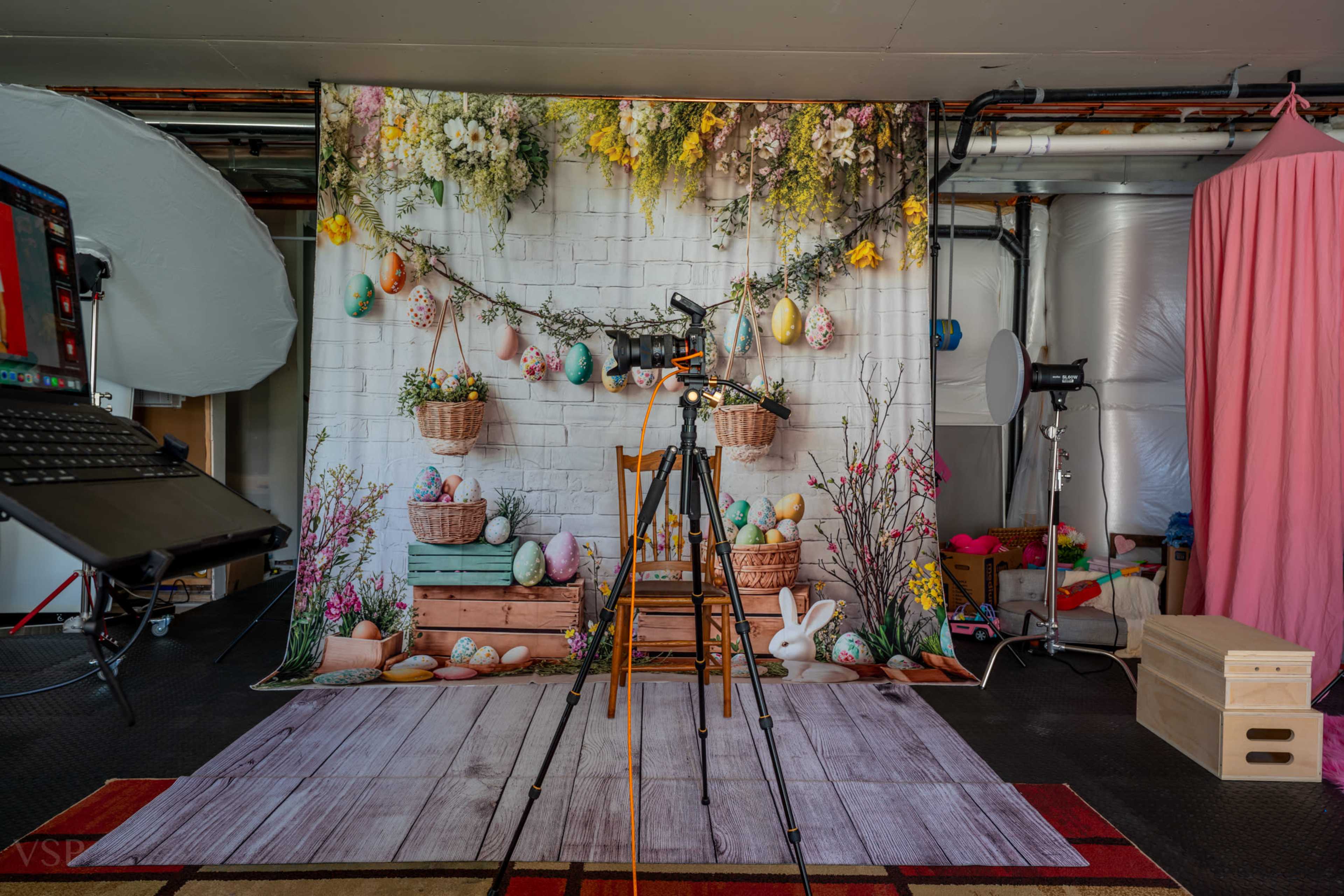 A photography setup features a floral backdrop decorated with Easter eggs, a chair, and various props, including a camera on a tripod.