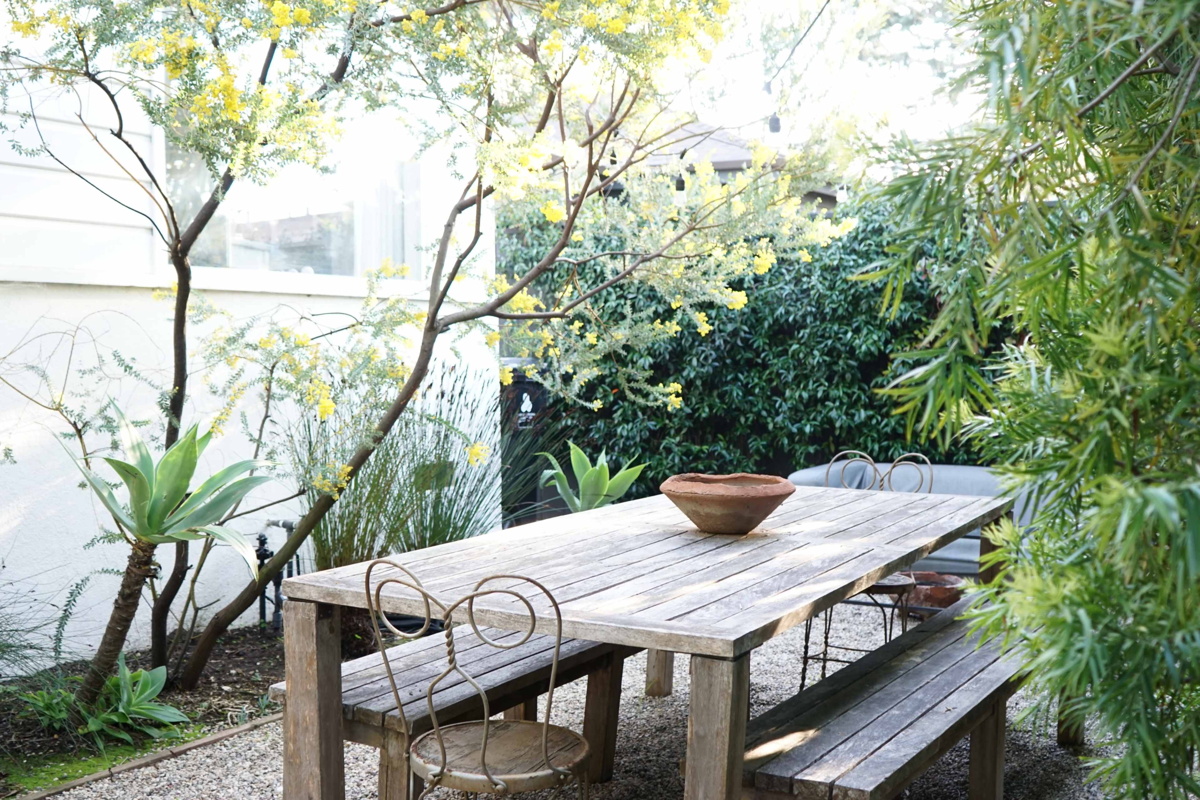 A wooden table and benches are set in a garden surrounded by flowering plants and trees.