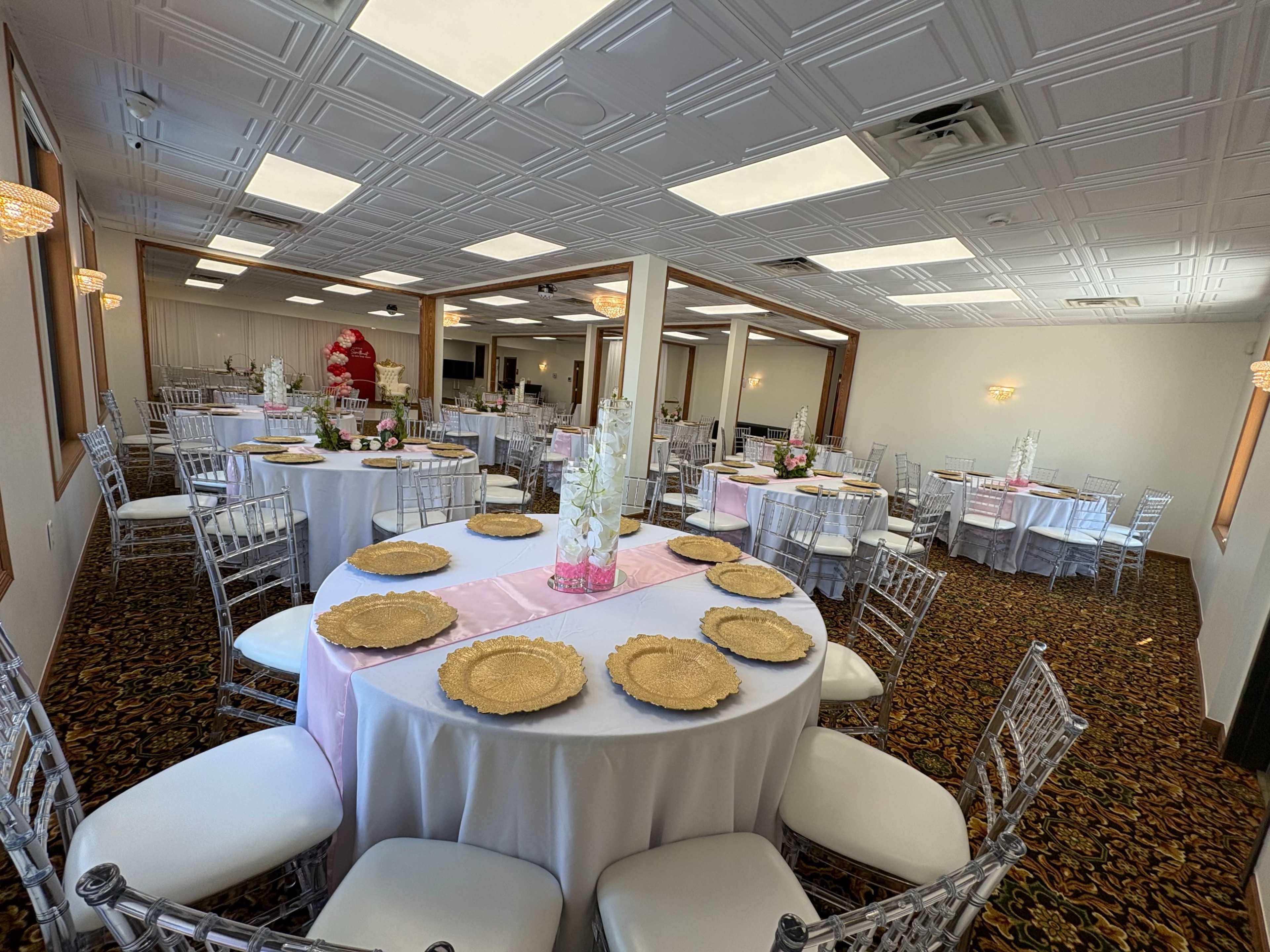 The image shows an event space set up with round tables covered in white tablecloths, gold chargers, and pink runners, surrounded by silver chiavari chairs.
