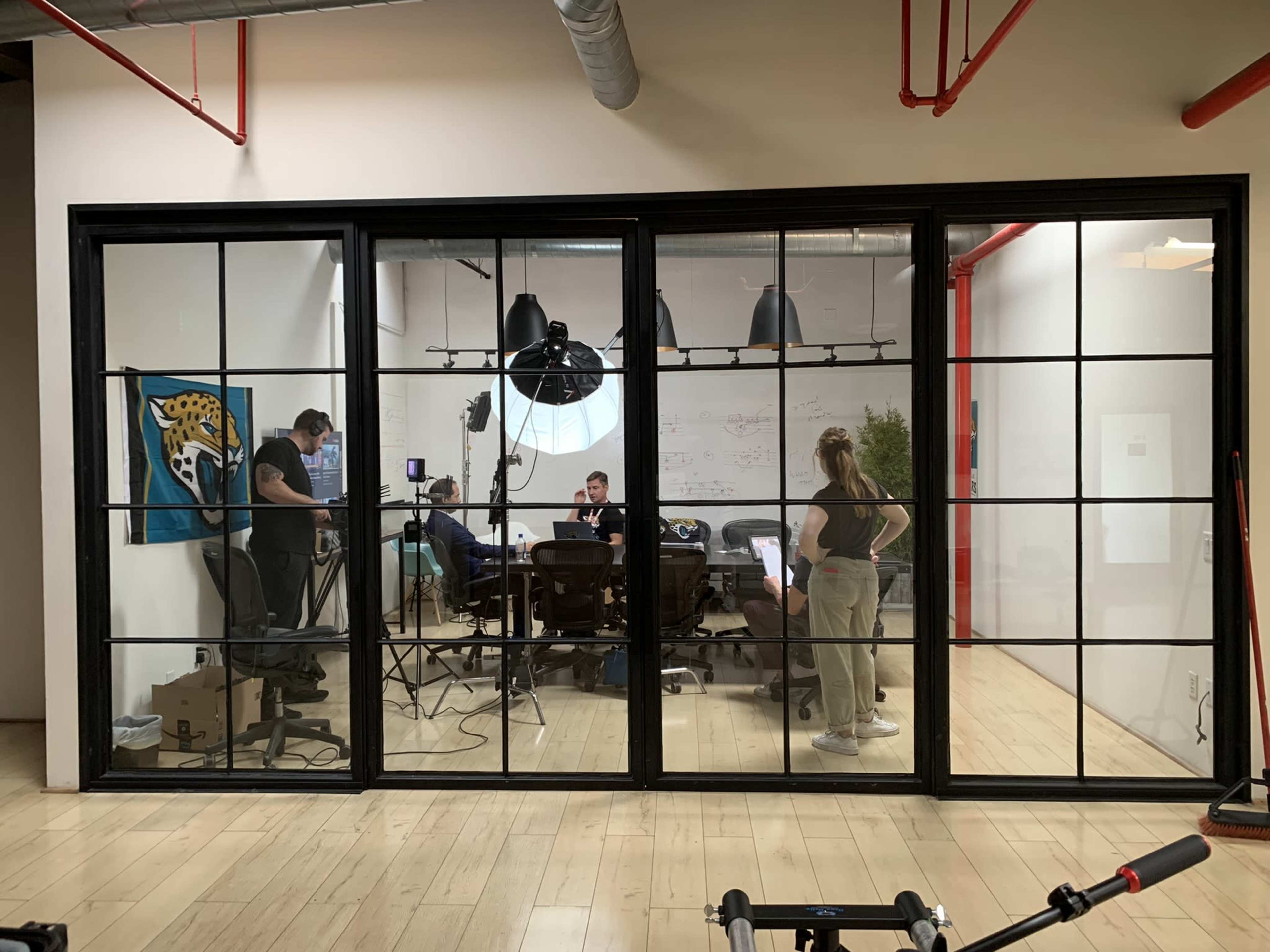 A group of people is working in a modern conference room, visible through large windows, with studio lighting equipment set up for a shoot.