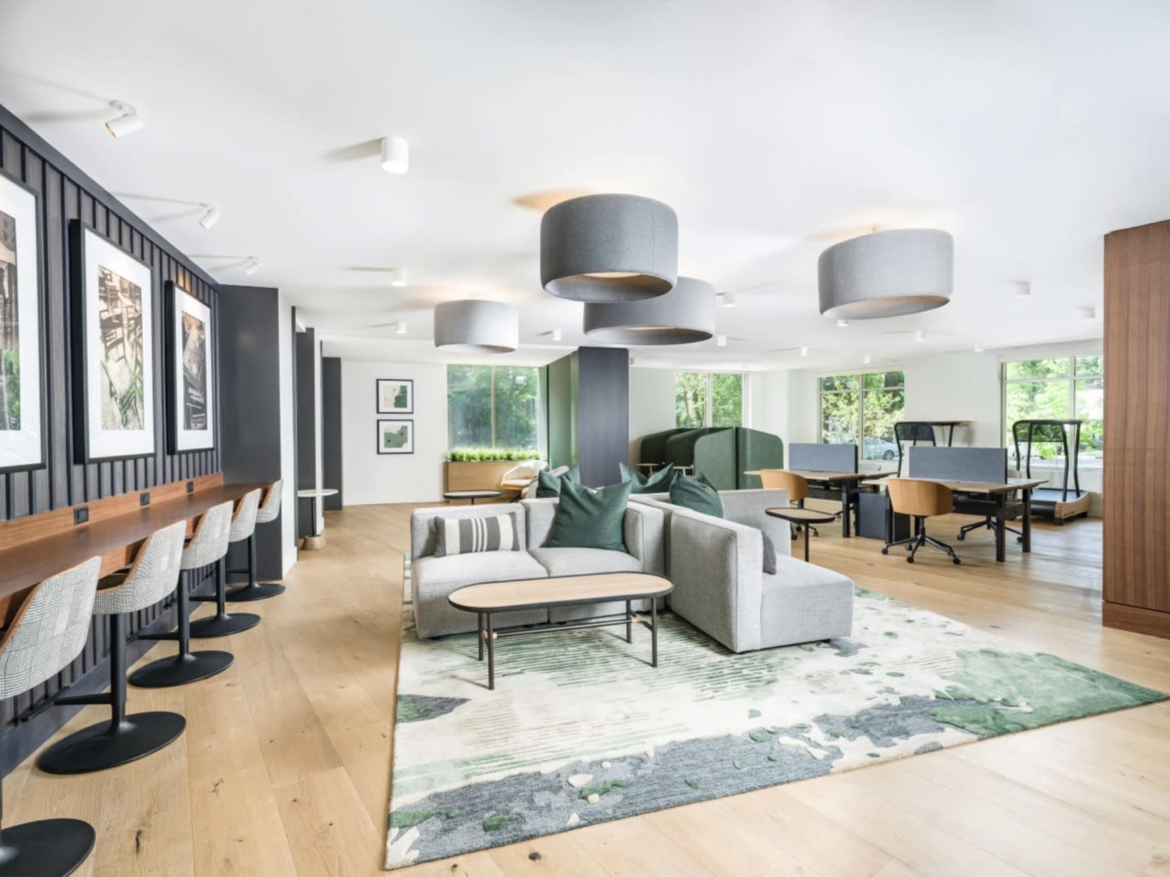 The image depicts a modern lounge area with a mix of seating options, including a gray sofa, circular tables, and bar stools, surrounded by large windows overlooking greenery.