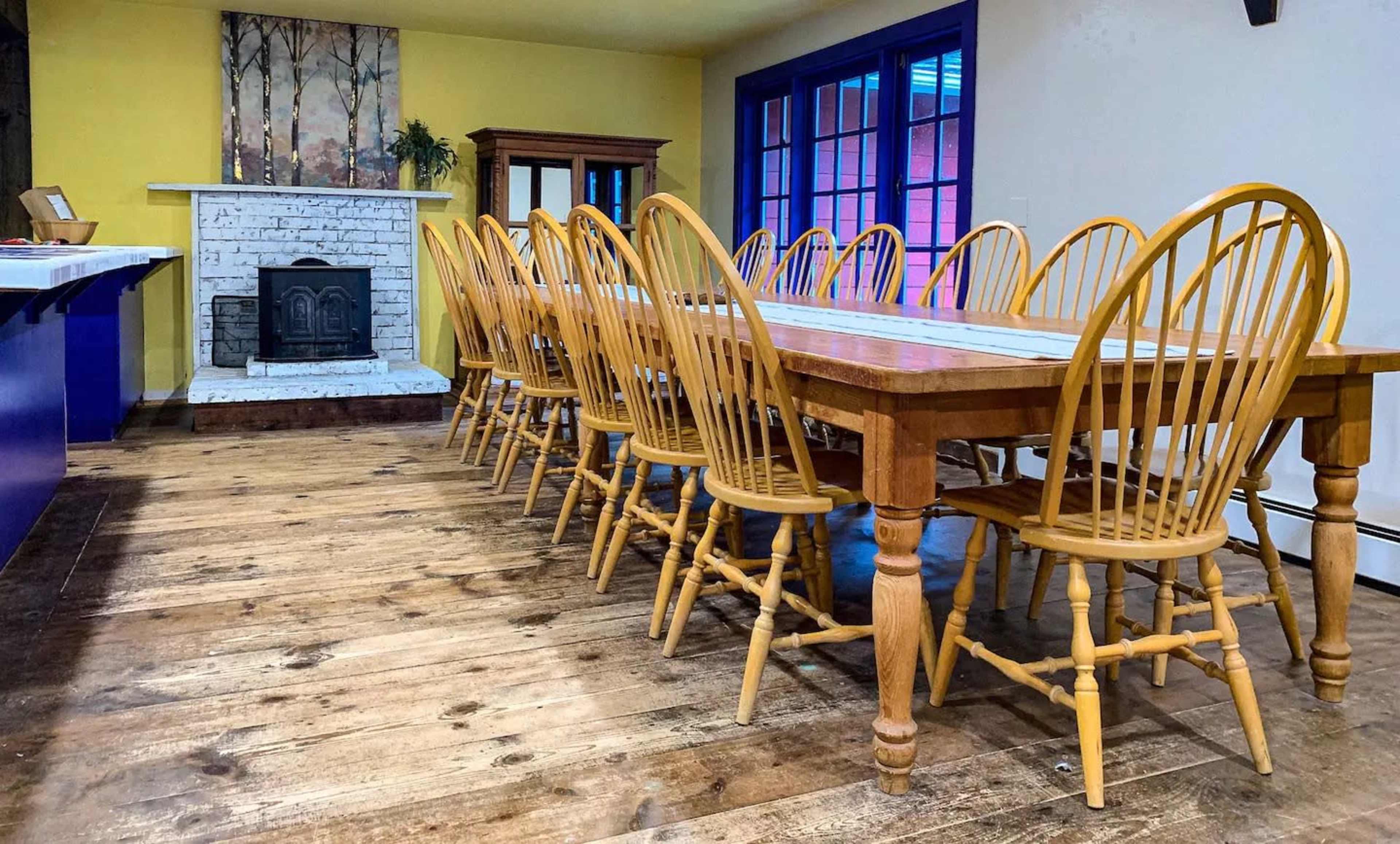 A long wooden dining table with numerous yellow chairs is set in a room featuring a white brick fireplace and colorful walls.