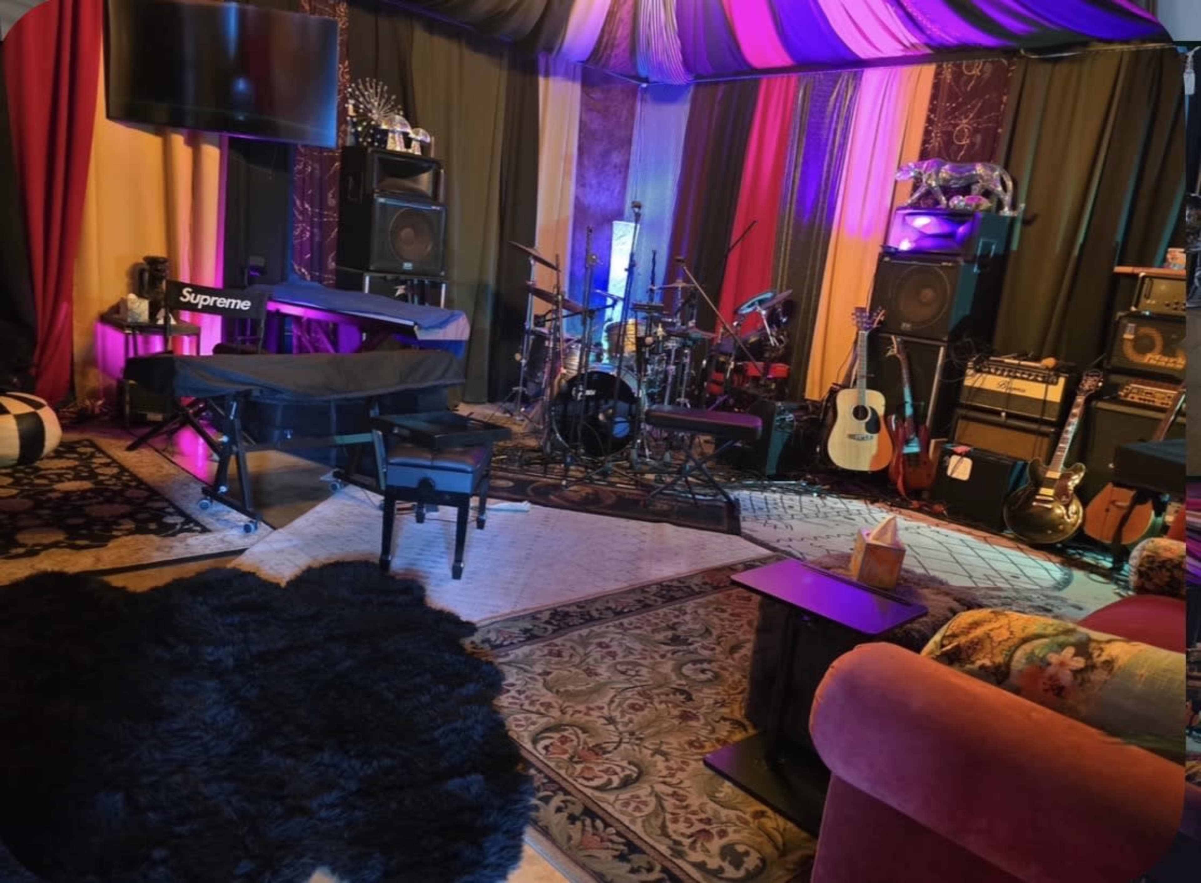 The image shows a colorful room filled with musical instruments, including a drum set, guitars, and amplifiers, alongside a piano and various seating options.