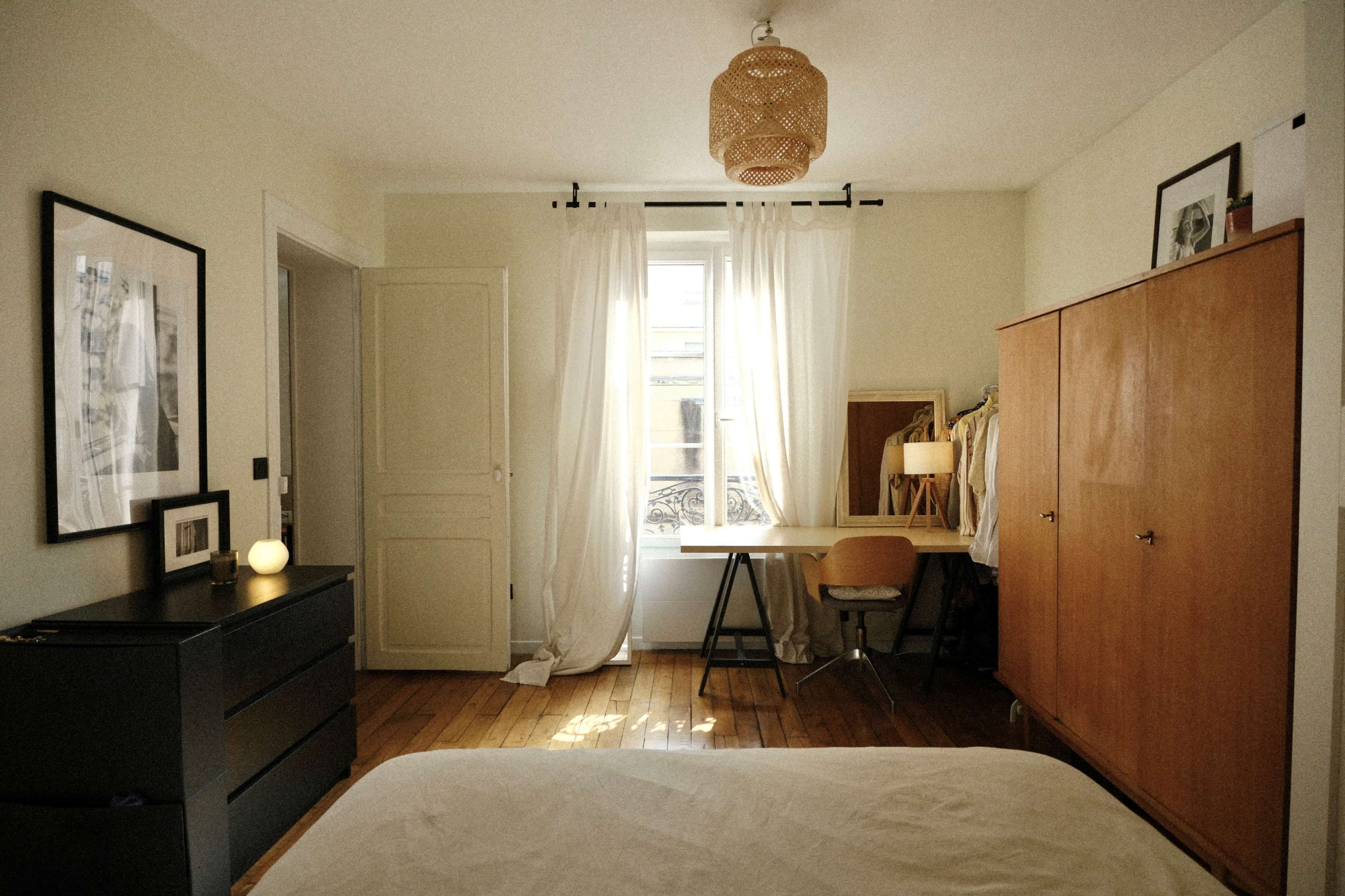 The image shows a bright bedroom featuring a bed in the foreground, a dresser, and a wooden wardrobe, with natural light coming through a window adorned with sheer curtains.
