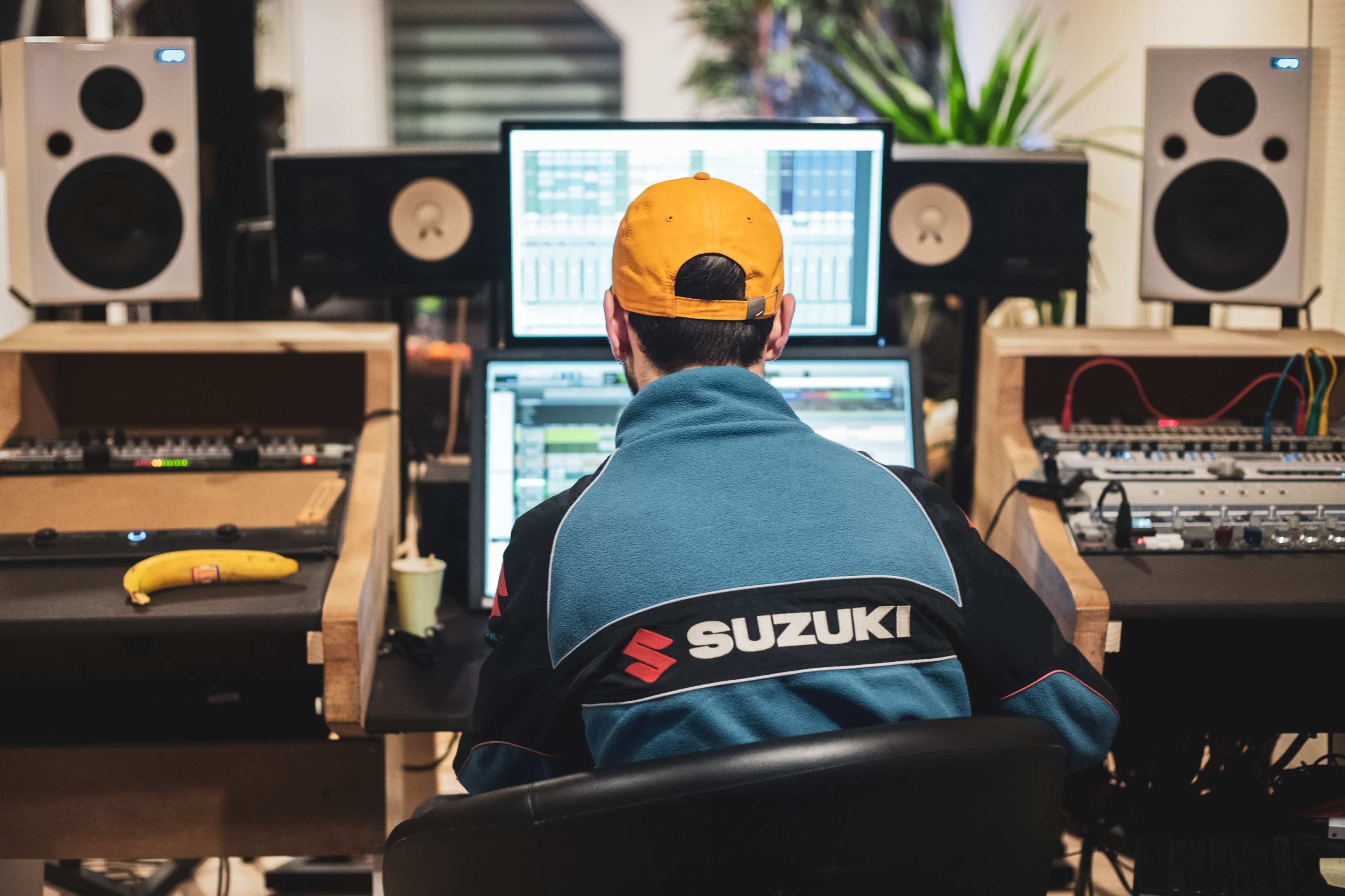 A person wearing a blue Suzuki jacket and yellow cap sits at a desk with multiple computer screens and audio equipment in a music studio.