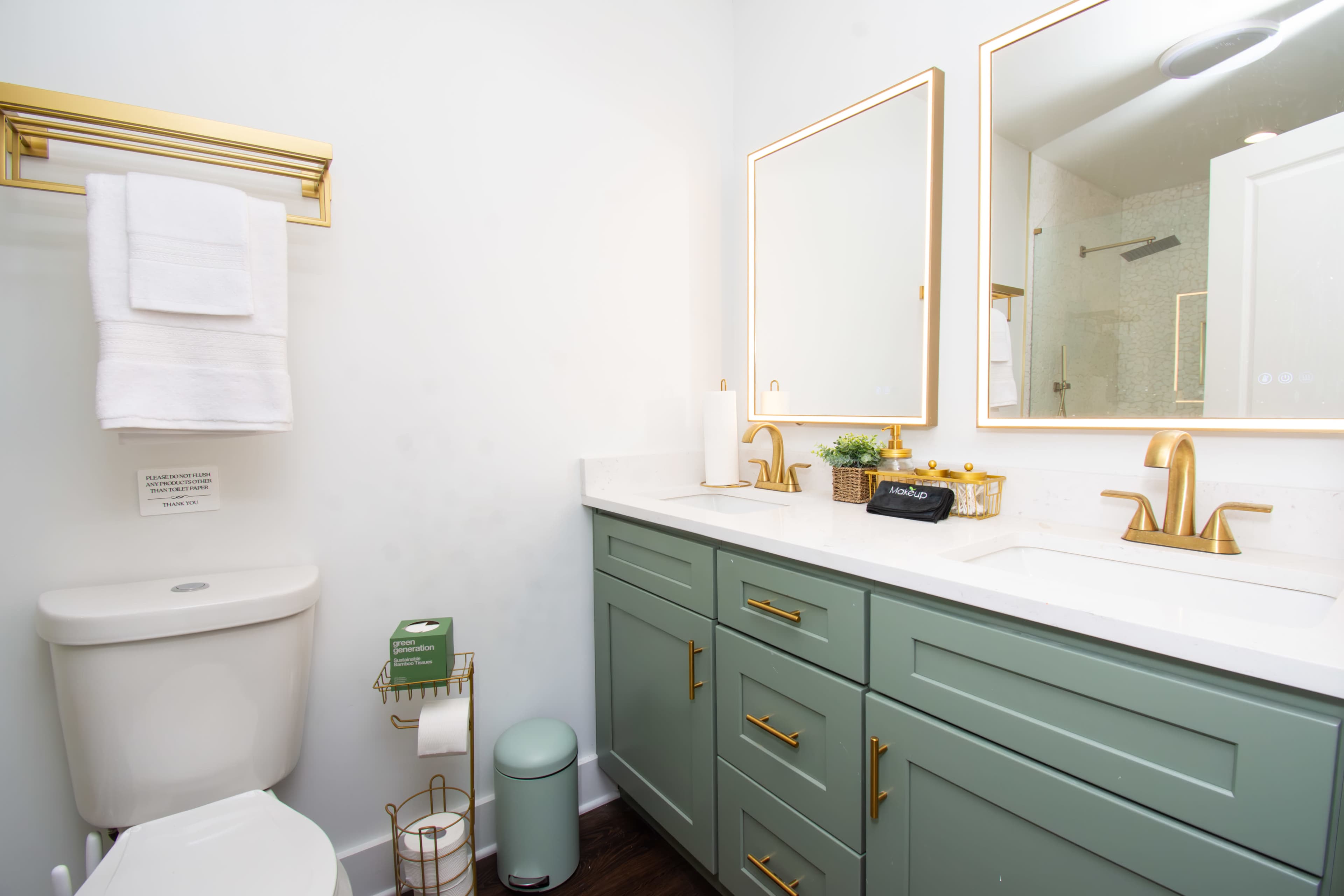 The image shows a modern bathroom featuring a double vanity with green cabinetry, two mirrors, a toilet, and a towel rack with white towels.