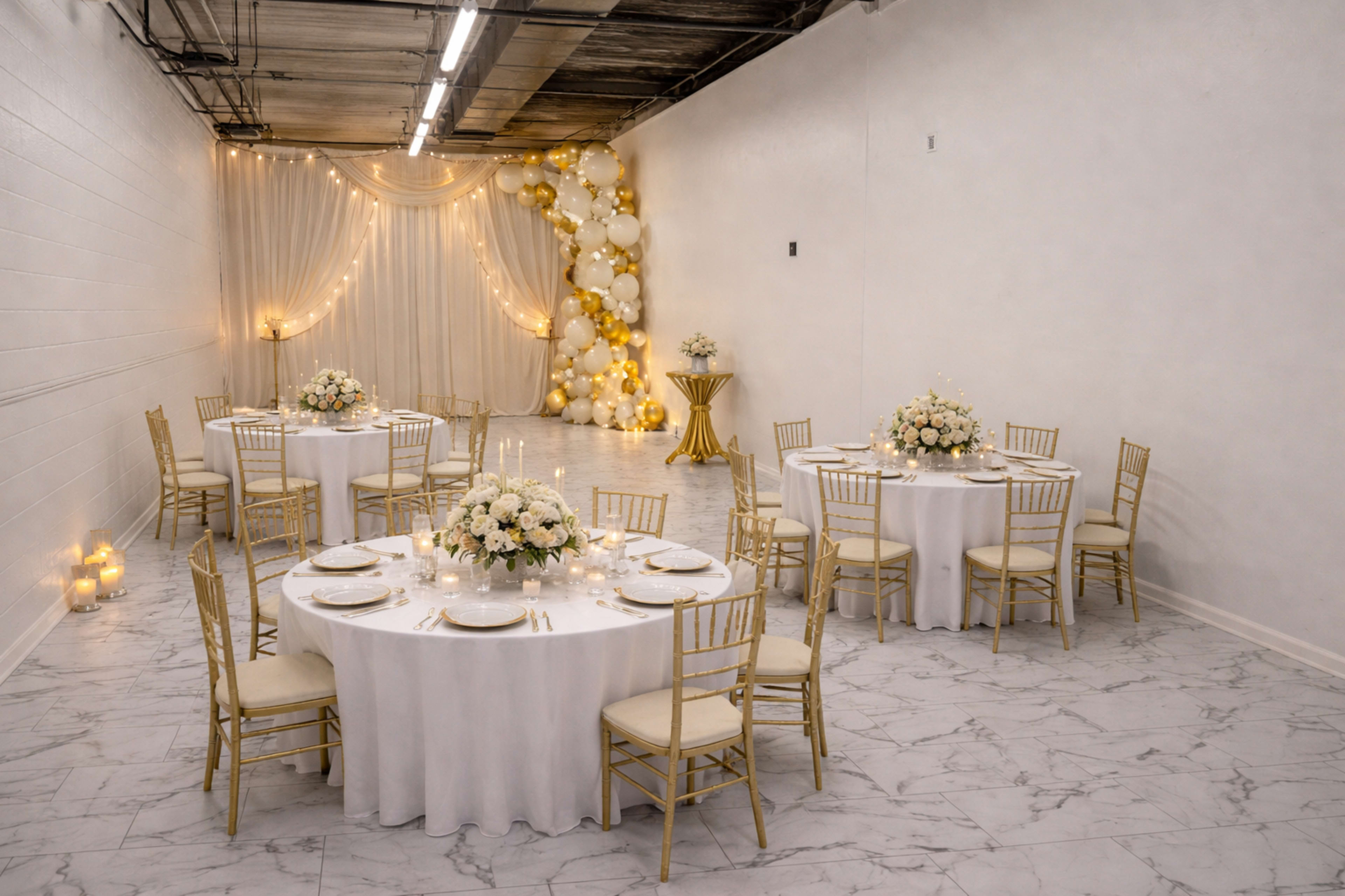 The image shows a spacious event venue featuring six elegantly set round tables with white tablecloths and gold chairs, surrounded by decorations of white and gold balloons, and soft lighting.