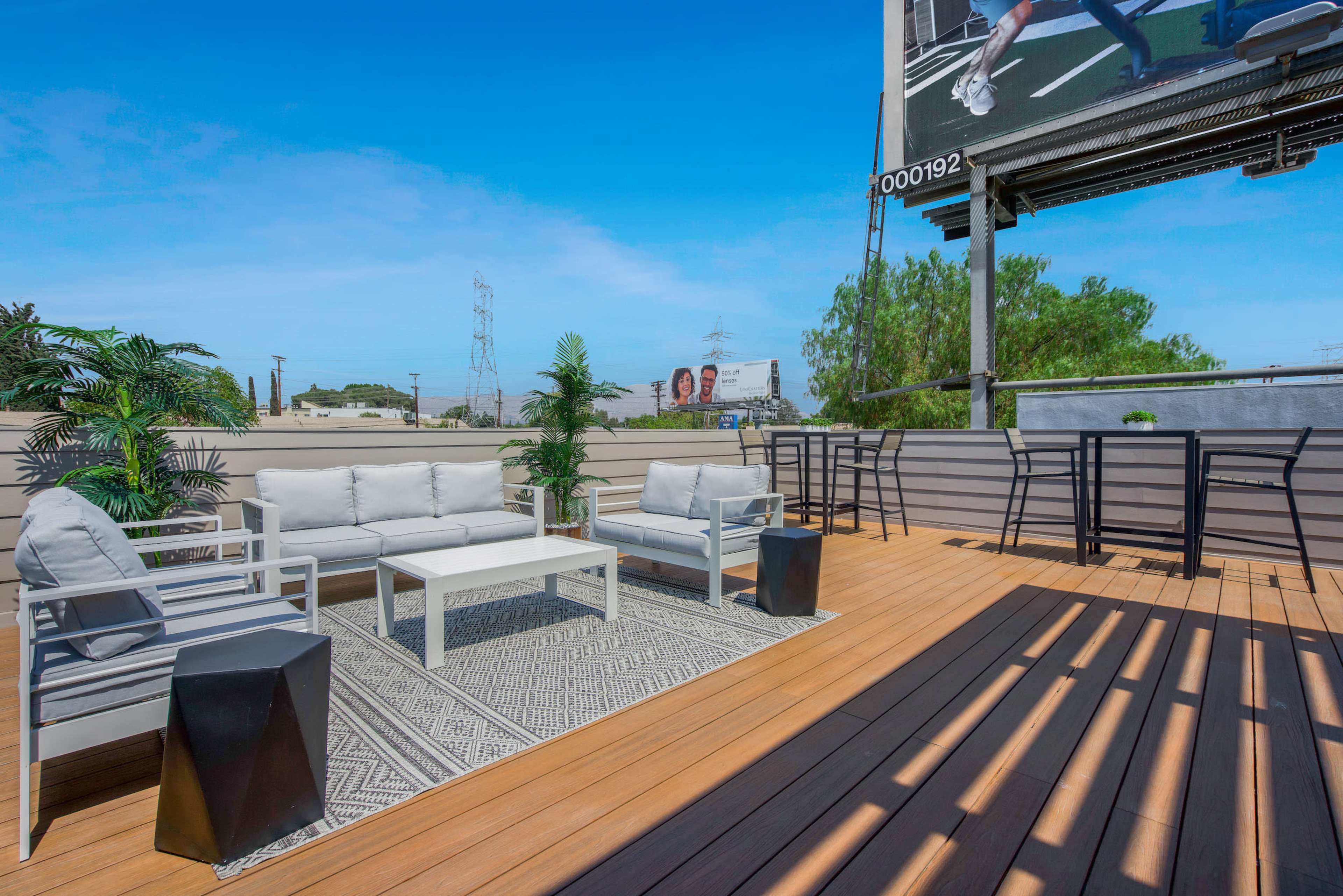 A modern outdoor seating area features a gray sofa set, geometric rug, and tall greenery, situated on a wooden deck beneath a blue sky.