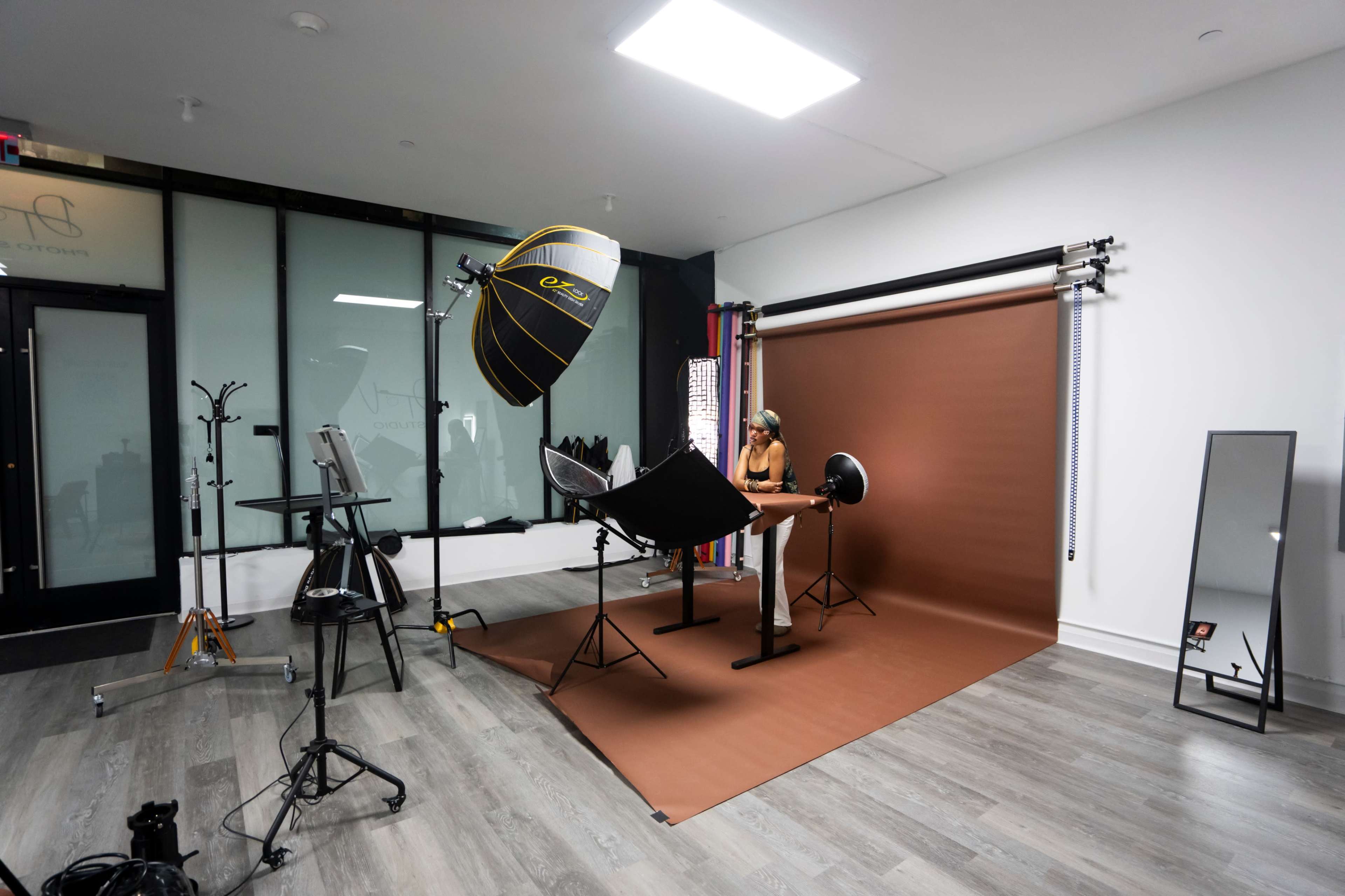 The image shows a photography studio setup with a model sitting at a table, surrounded by lighting equipment and a backdrop.