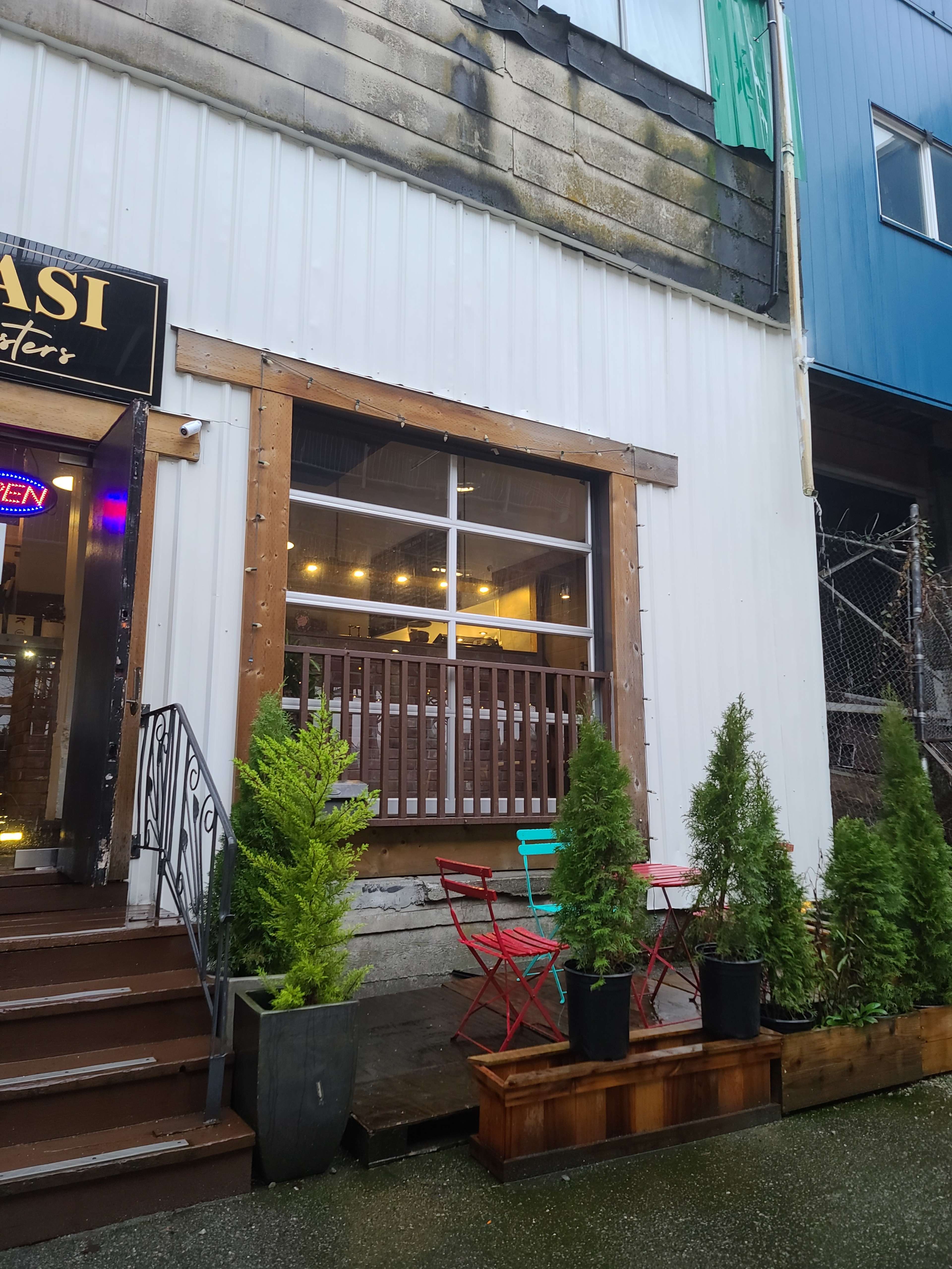 The image shows a café entrance with a wooden frame, flanked by two small evergreen shrubs, and featuring colorful outdoor seating with red and blue chairs.