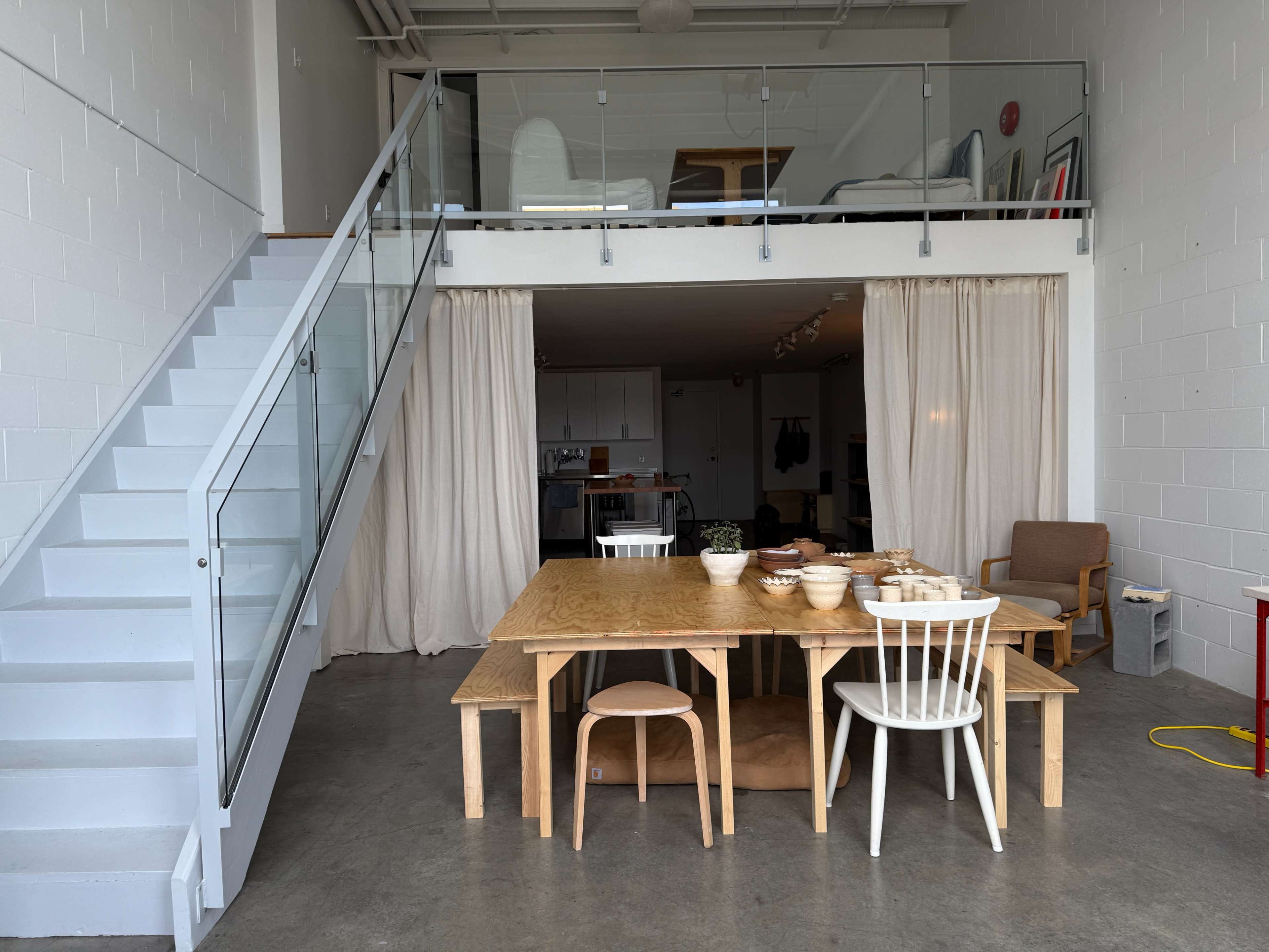 A wooden dining table surrounded by chairs is set in a spacious, minimalist interior with a staircase leading to a loft area.