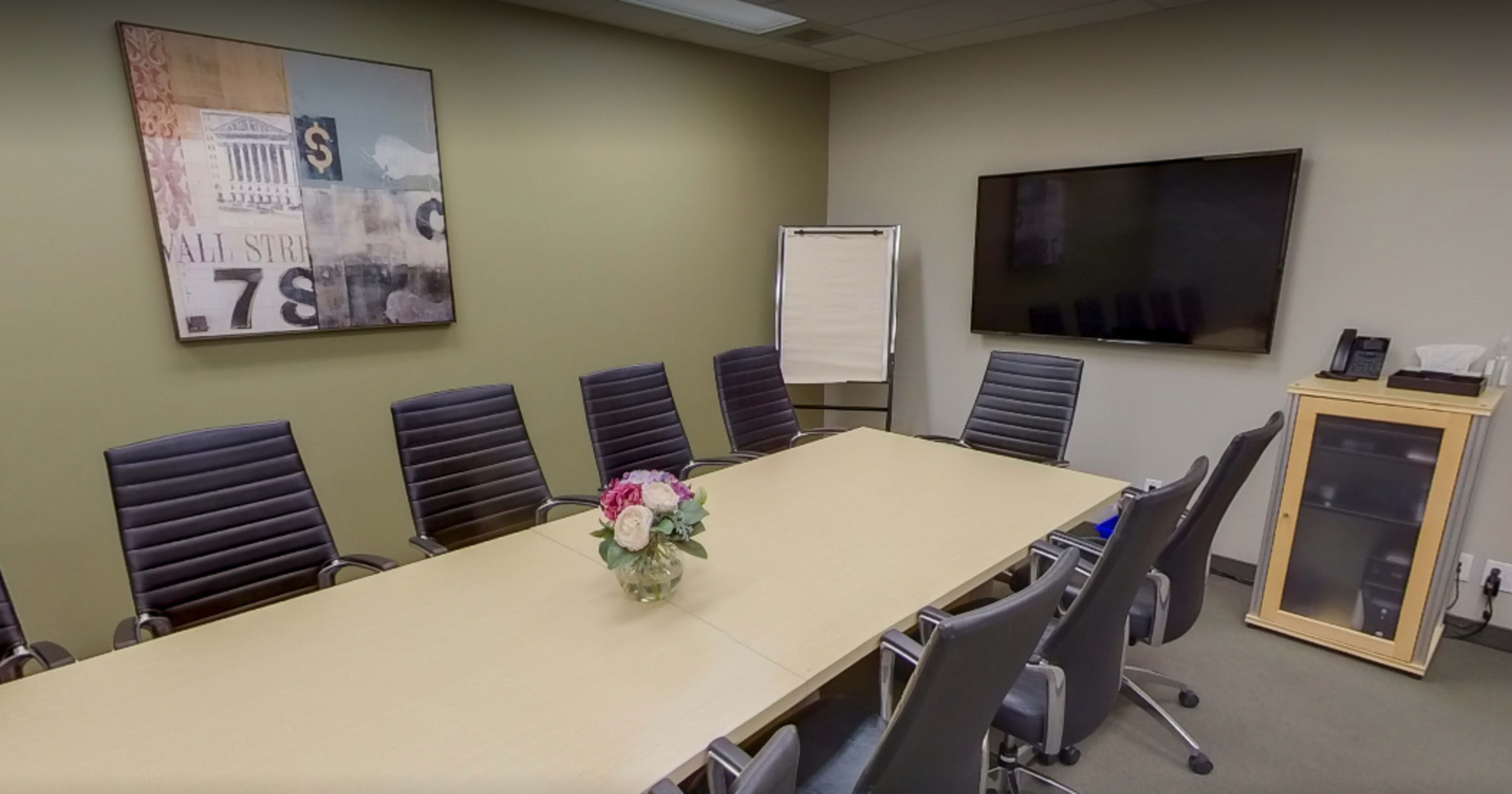 A conference room features a long table surrounded by several black chairs, a flower arrangement in the center, and a large screen mounted on the wall.