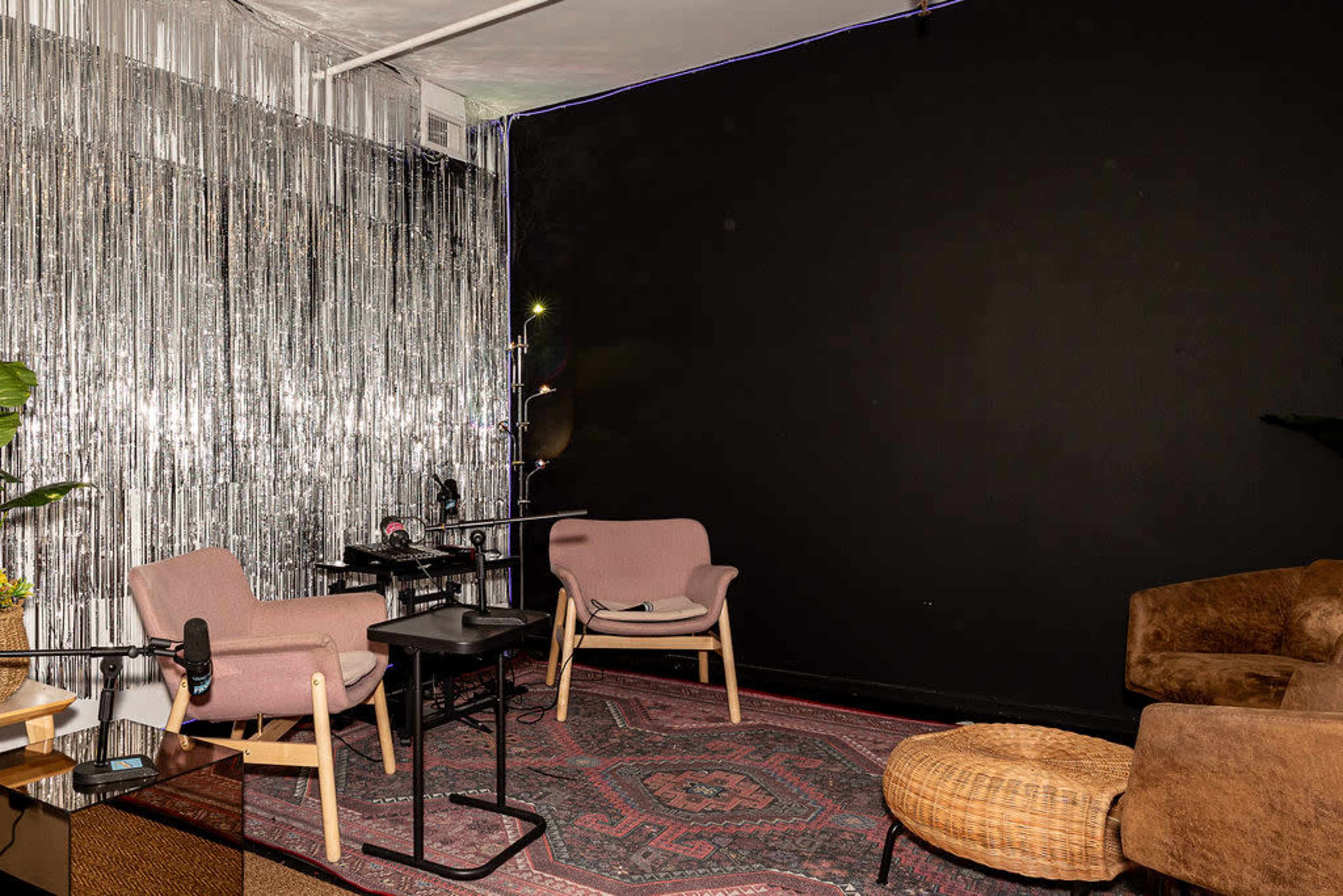 The image shows a cozy room with two pink chairs, a small table, and a wall draped in metallic fringe near a dark-colored wall.