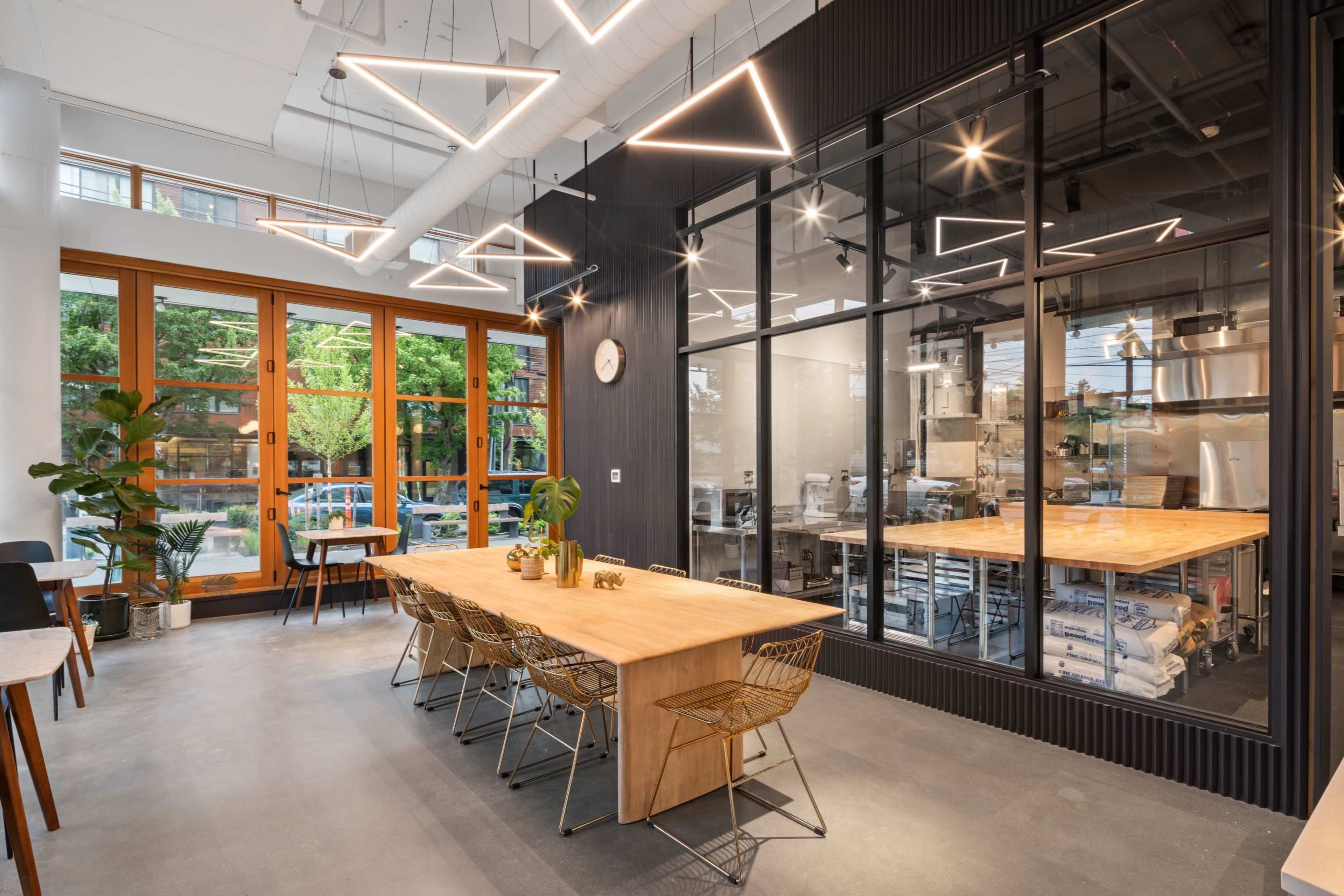 A modern workspace features a long wooden table surrounded by metal chairs, with large windows and a glass-walled kitchen area visible in the background.