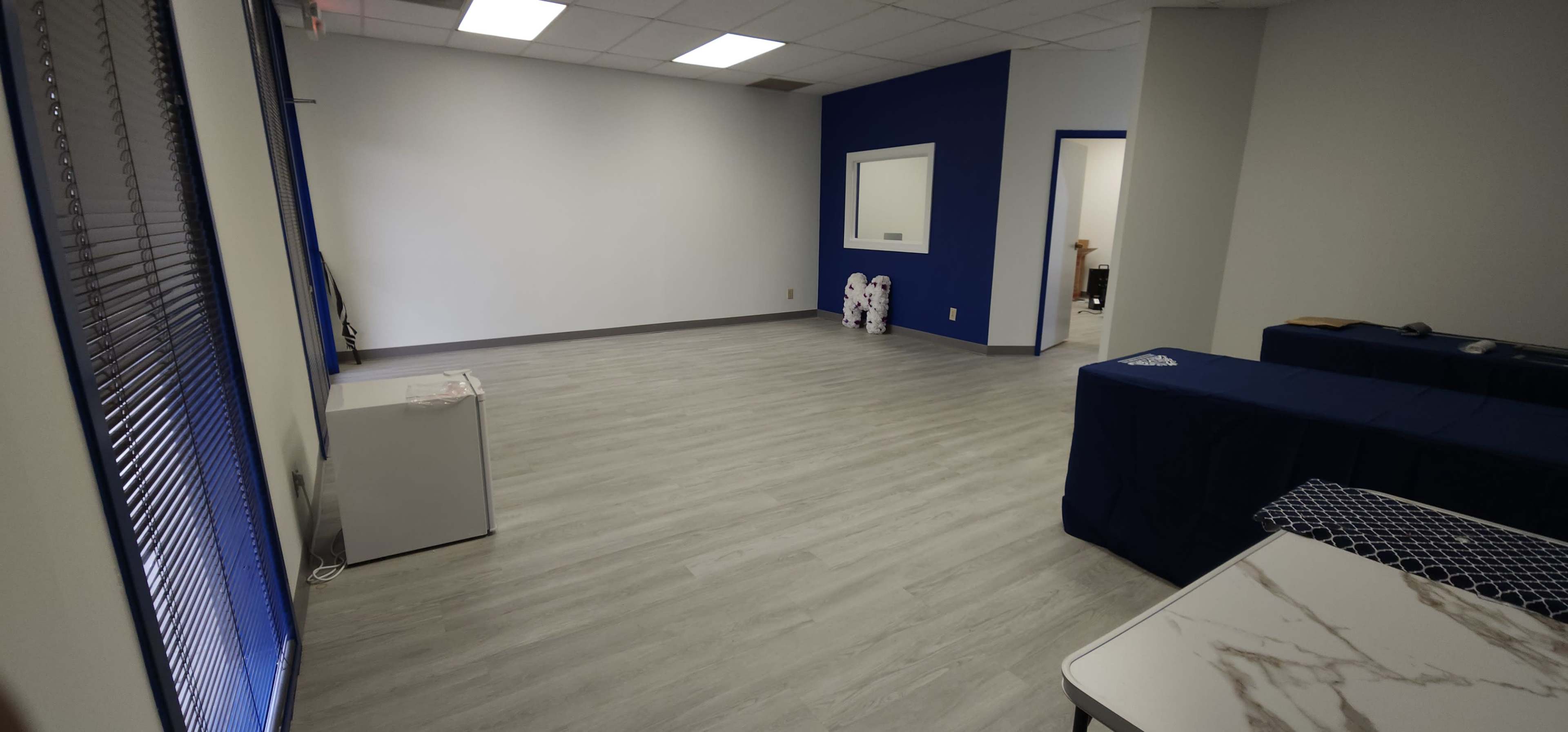 The image shows a spacious, uncluttered room with light-colored flooring, a blue accent wall, and a few pieces of furniture near the corners.