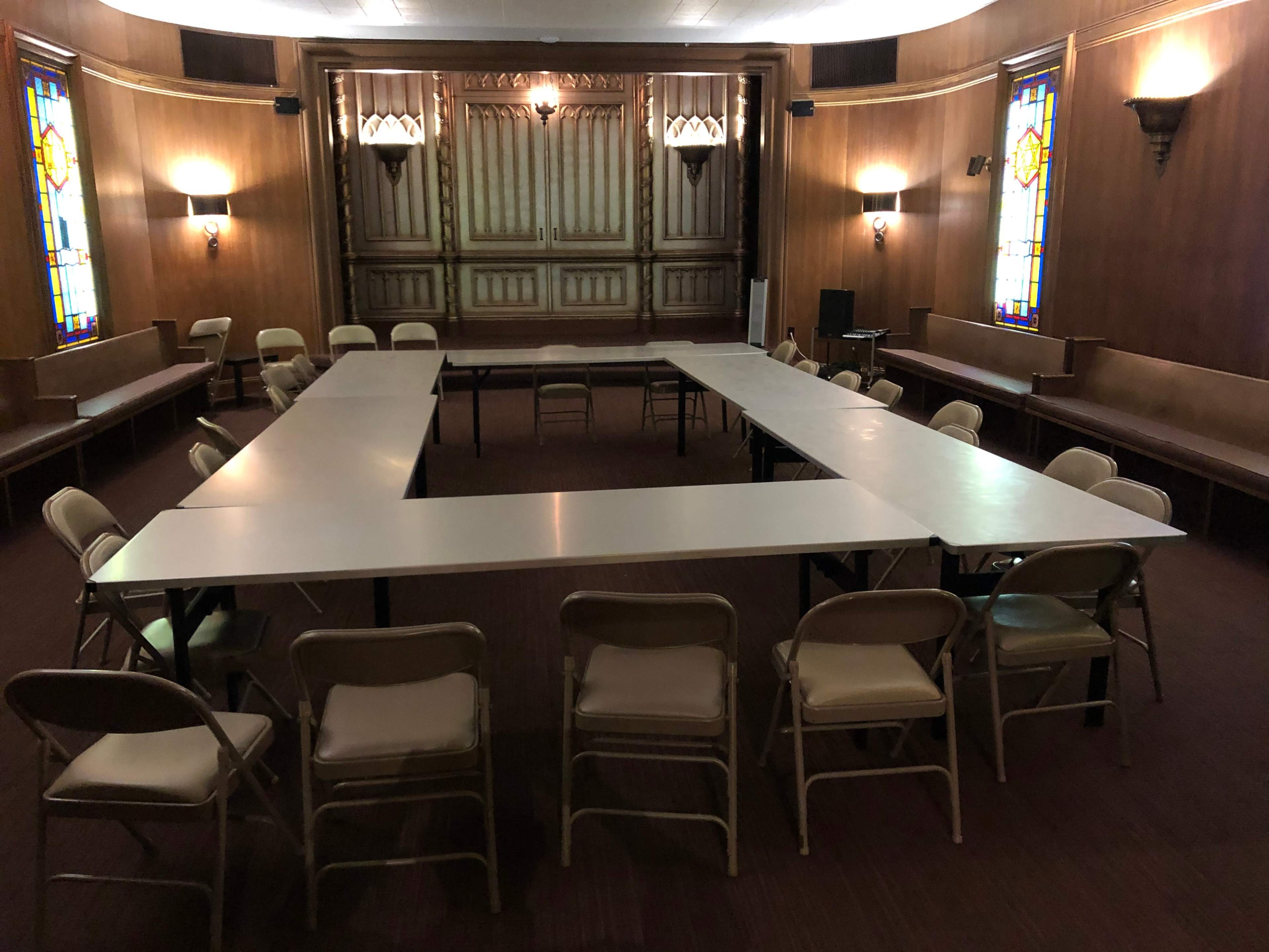 The room features a U-shaped arrangement of tables with chairs surrounding it, set against a backdrop of wooden paneling and stained glass windows.