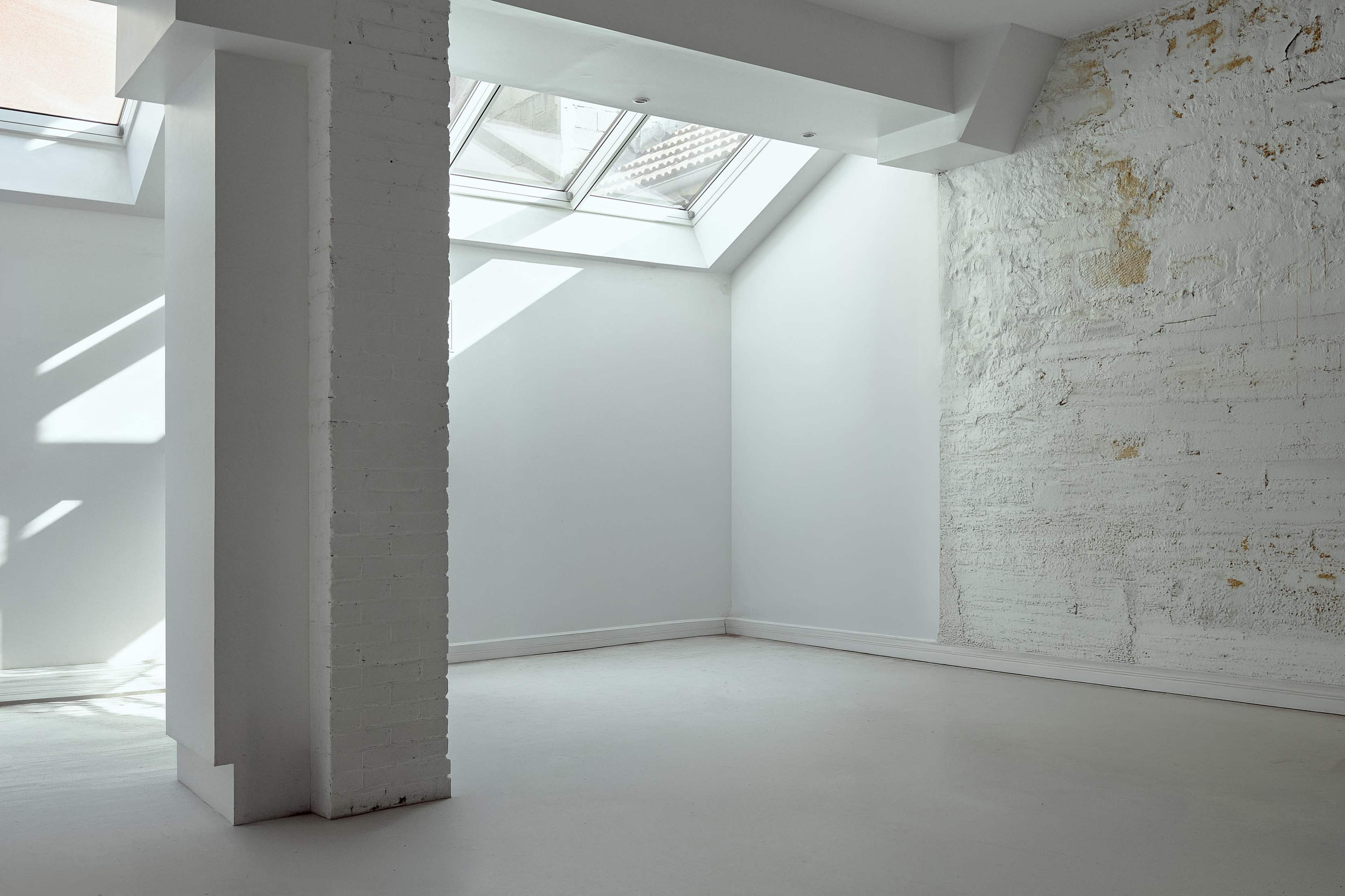 The image shows a minimalist interior space with a skylight and one exposed brick wall.