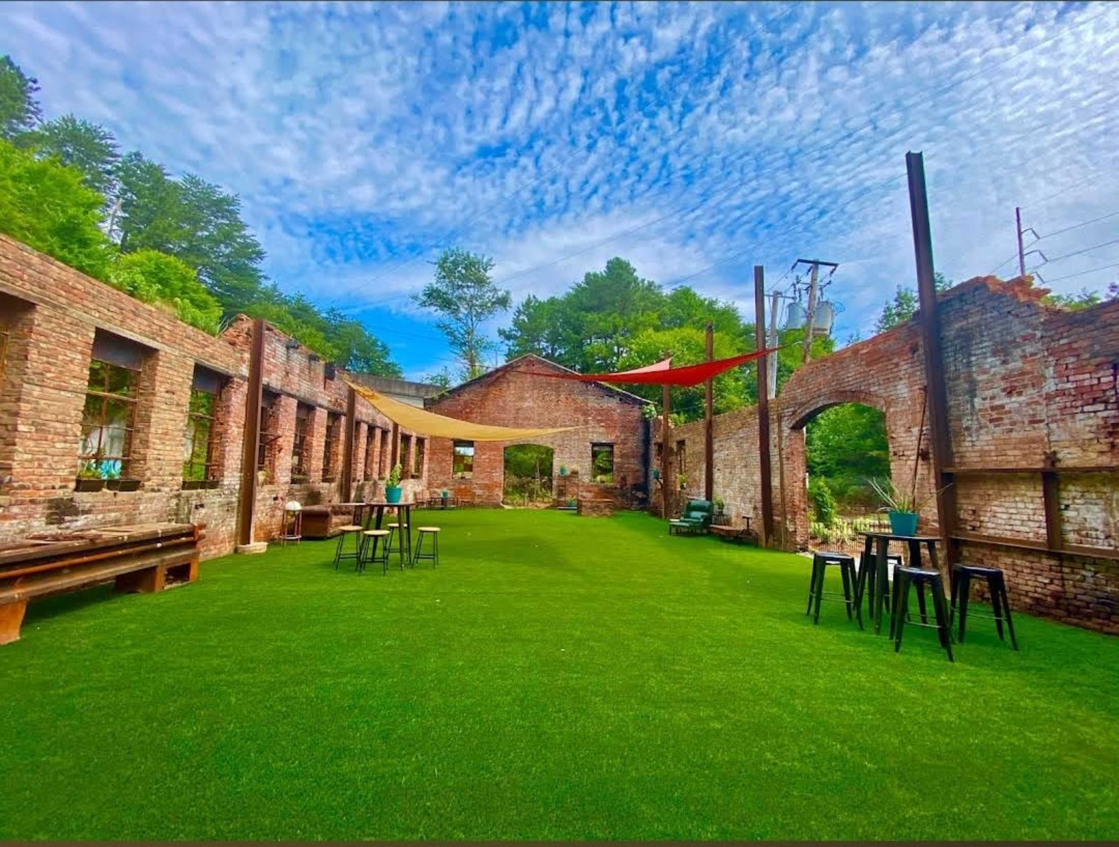 The image shows a vibrant green lawn surrounded by the ruins of a brick building with large openings and remnants of walls, under a blue sky with scattered clouds.