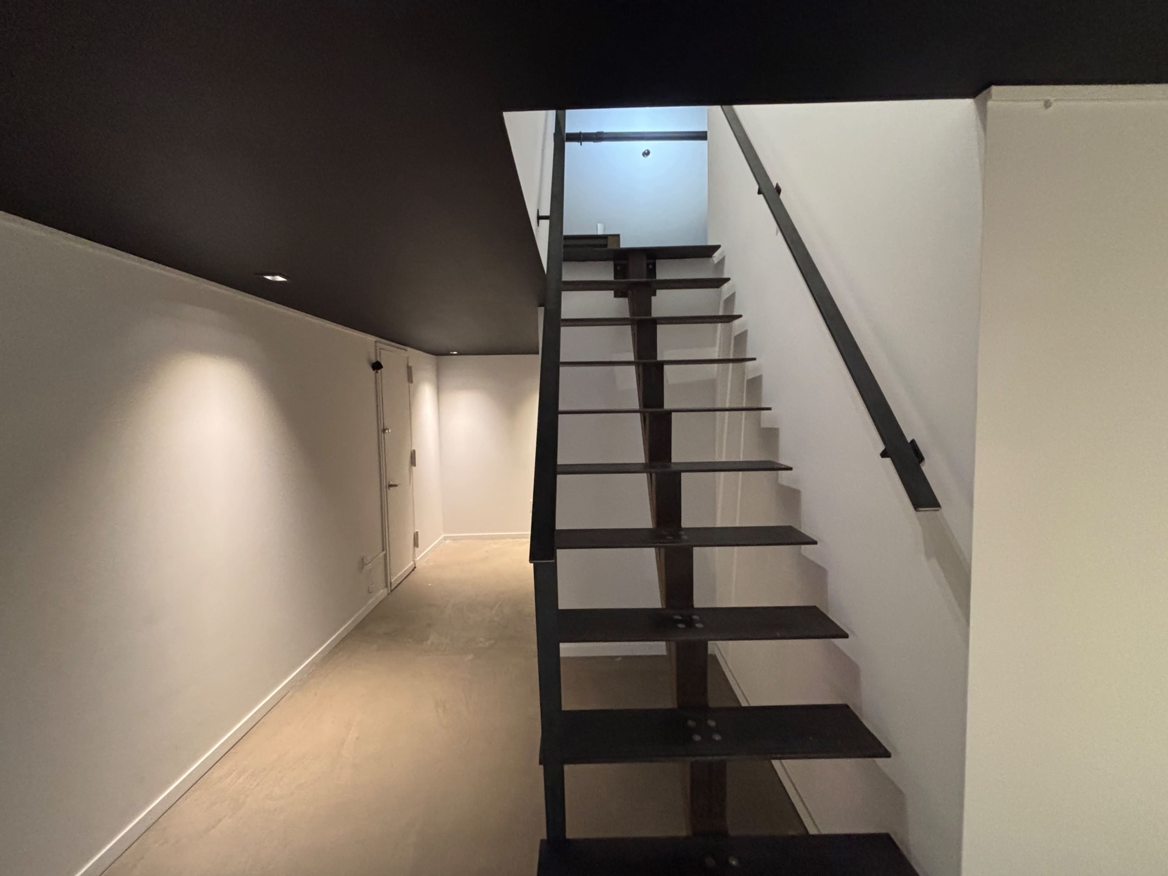 The image shows a modern staircase with black metal steps leading to an upper level, set in a minimalist interior with white walls and a dark ceiling.