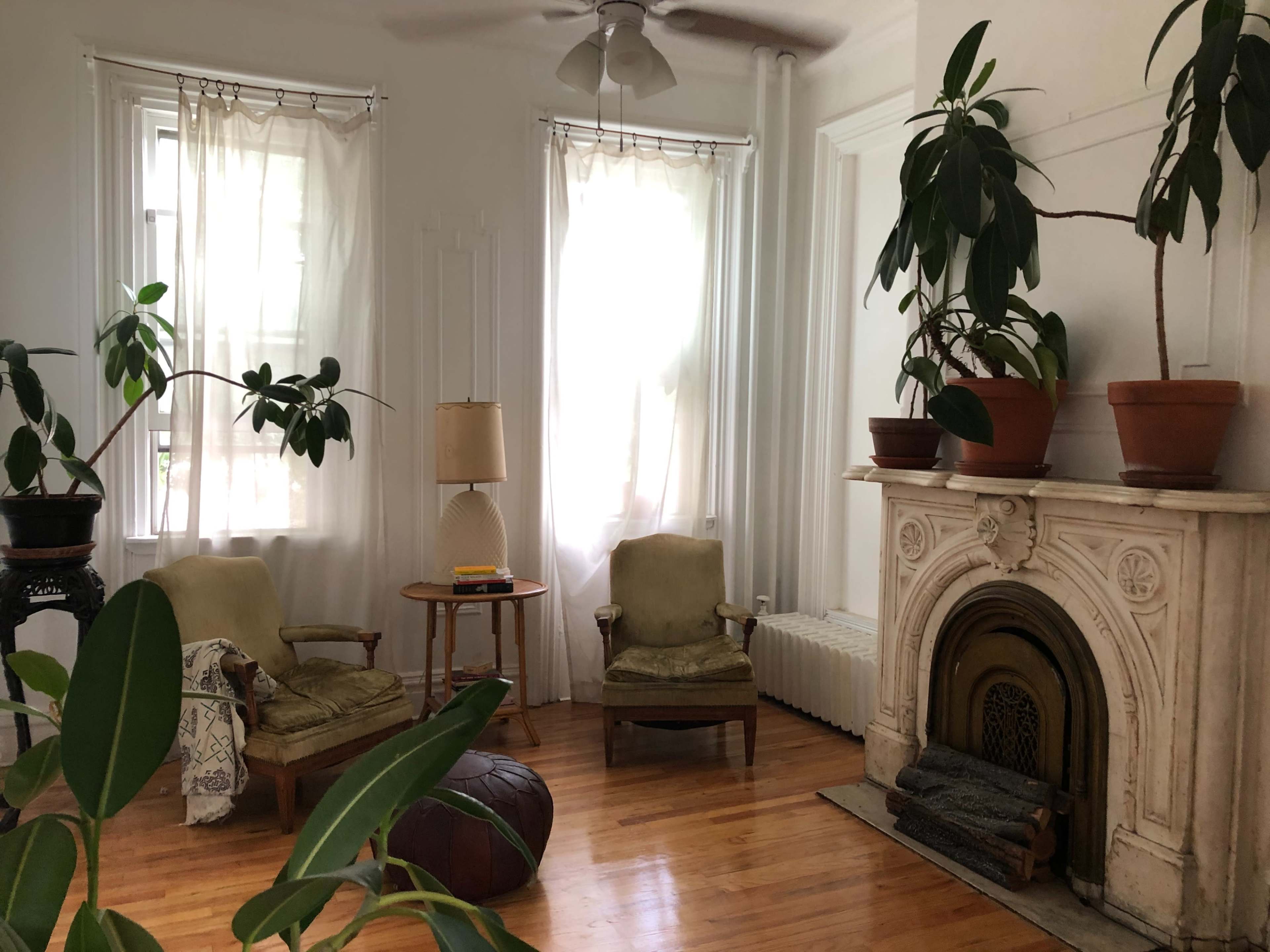 A bright living room features two chairs, a lamp, a piano, and a decorative fireplace surrounded by potted plants.