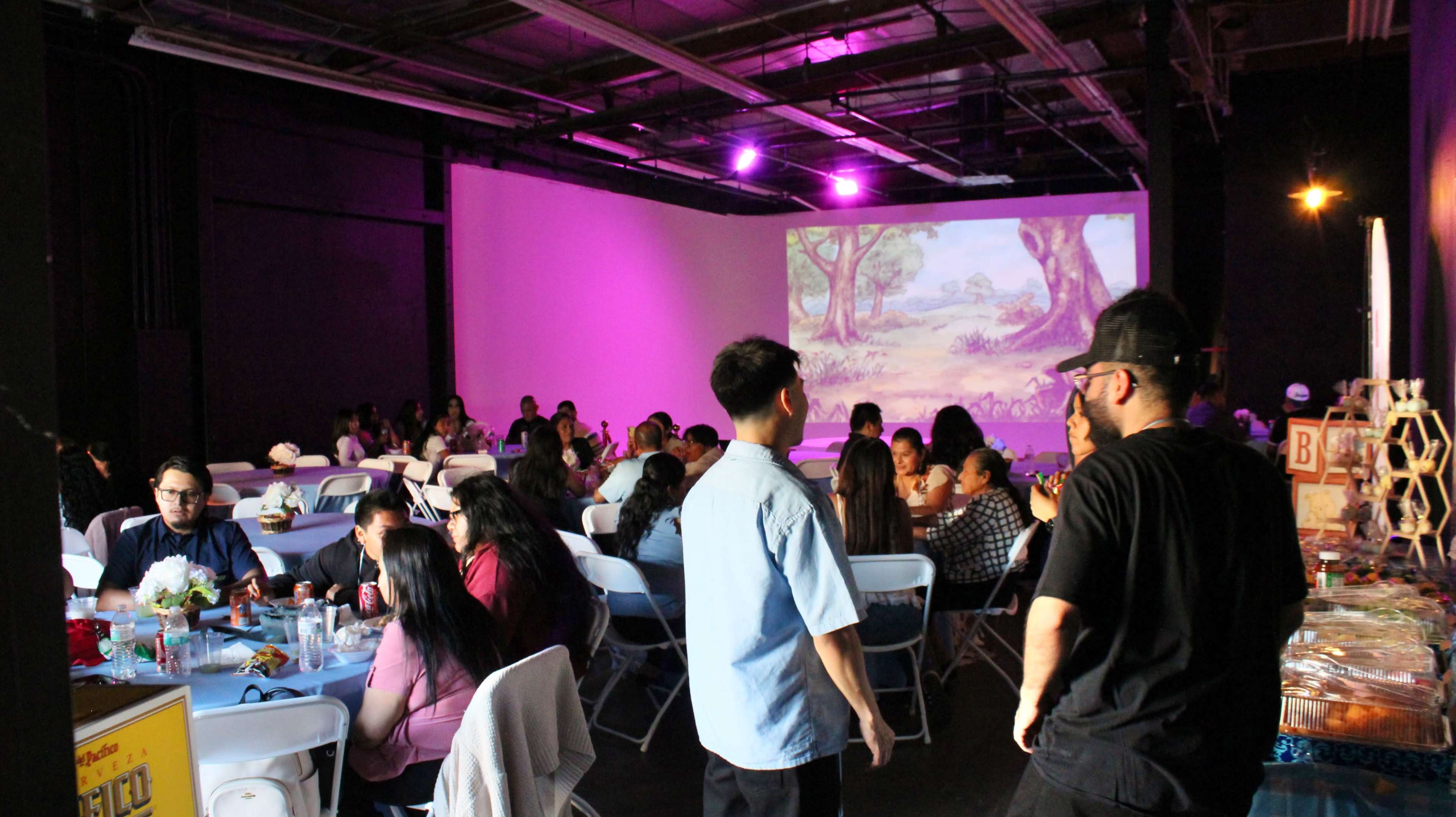 A gathering of people seated at tables with a large screen displaying an animated scene in a dimly lit venue.