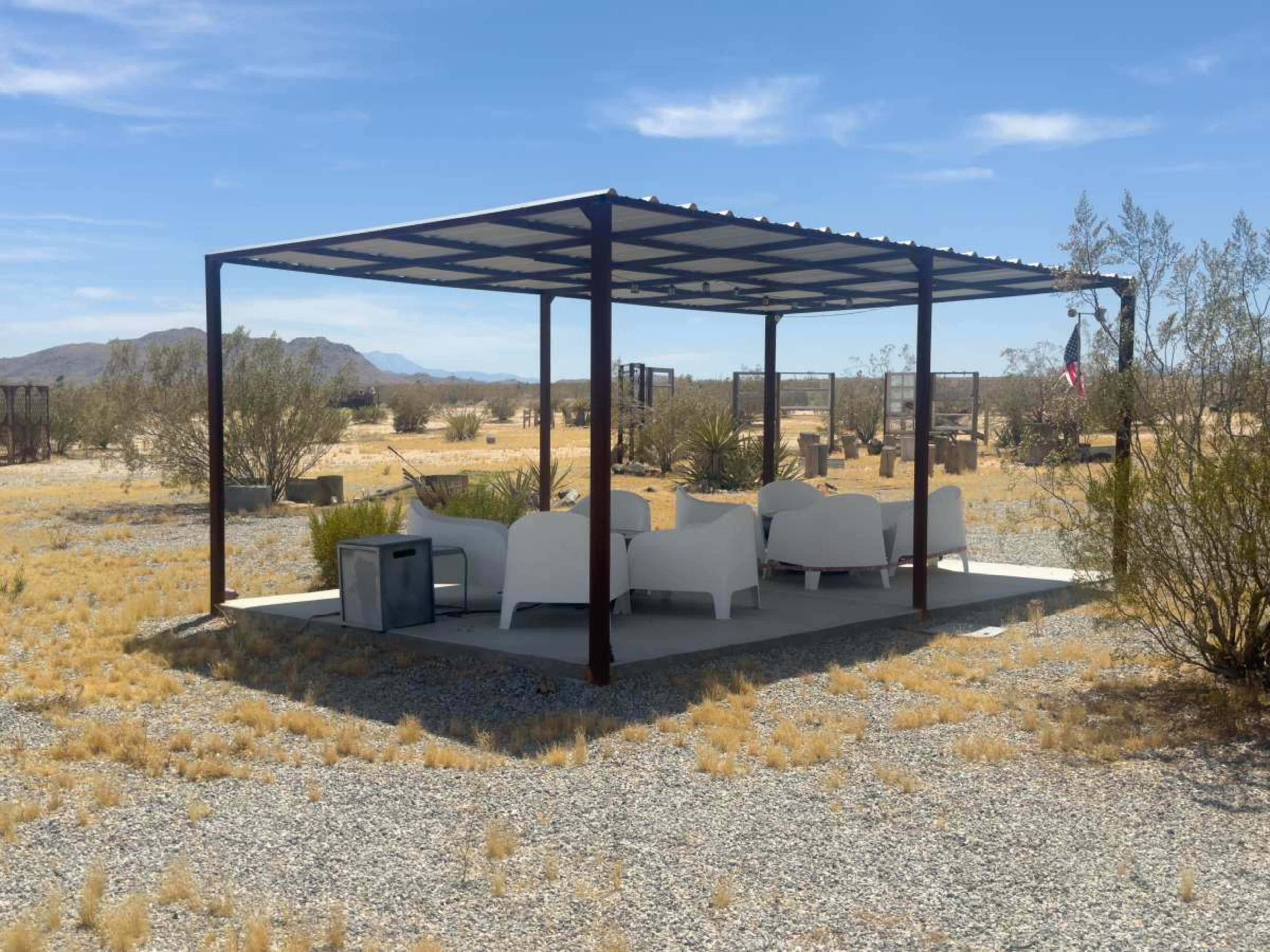 A shaded seating area with white furniture is situated on a gravel surface in a desert landscape.
