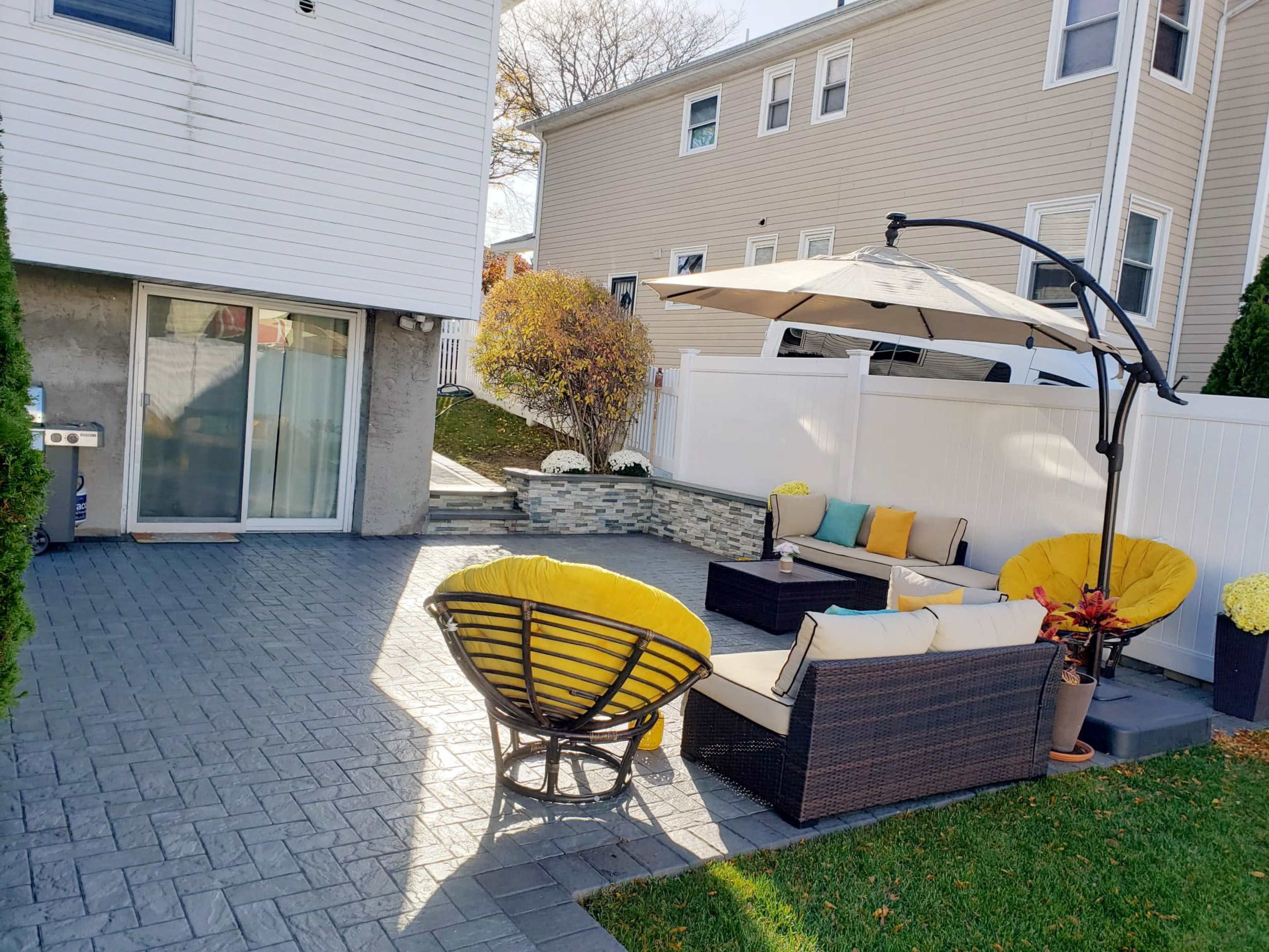 The image shows a landscaped outdoor seating area with a patio featuring a dining set, lounge chairs, and an umbrella beside a house.
