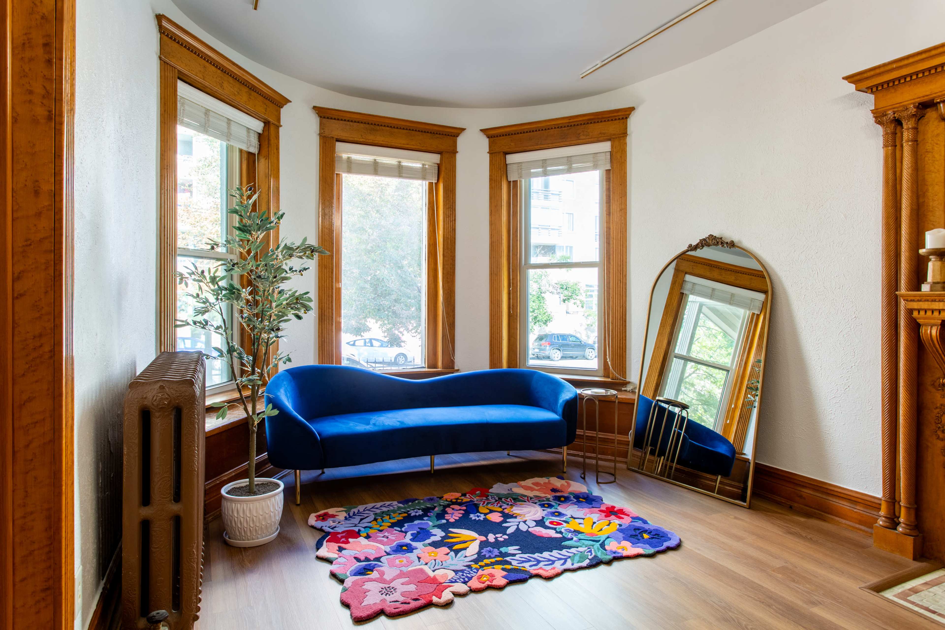 A blue sofa is positioned against a wall near large windows, with a decorative mirror and a colorful rug on the wooden floor.
