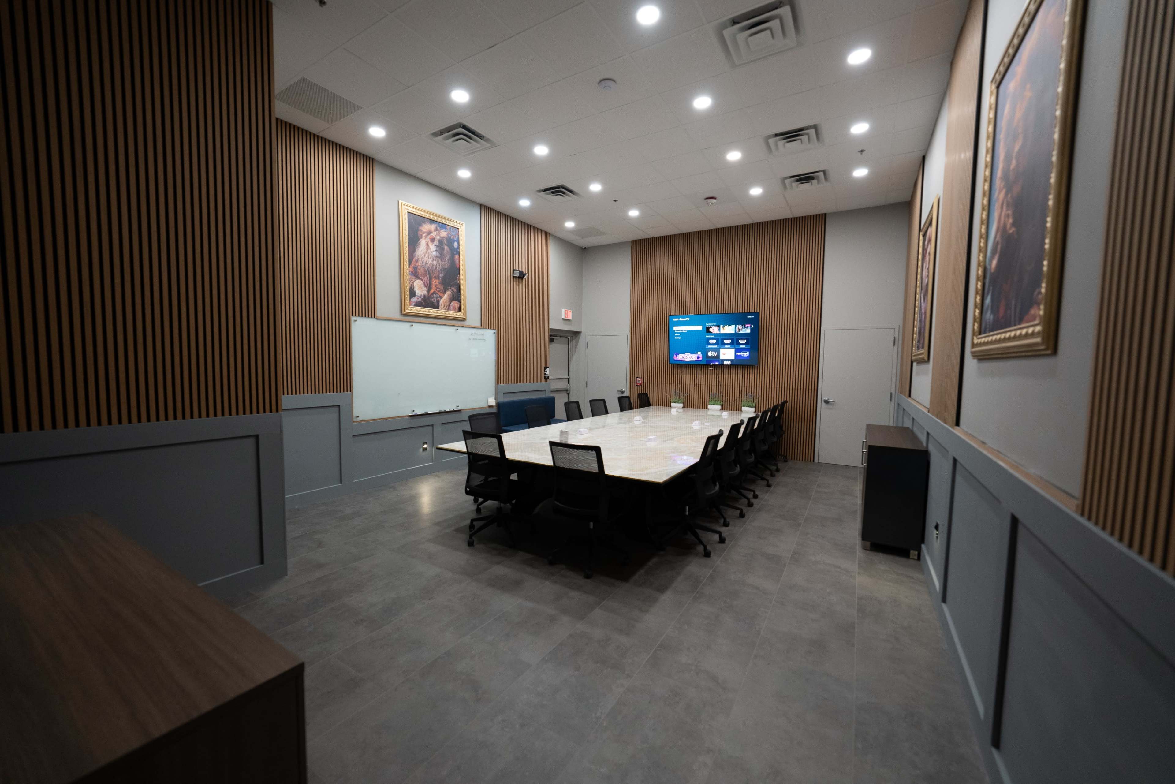The image shows a large conference room with a long table, surrounded by black chairs, and features a whiteboard, a TV display, and framed artwork on the walls.