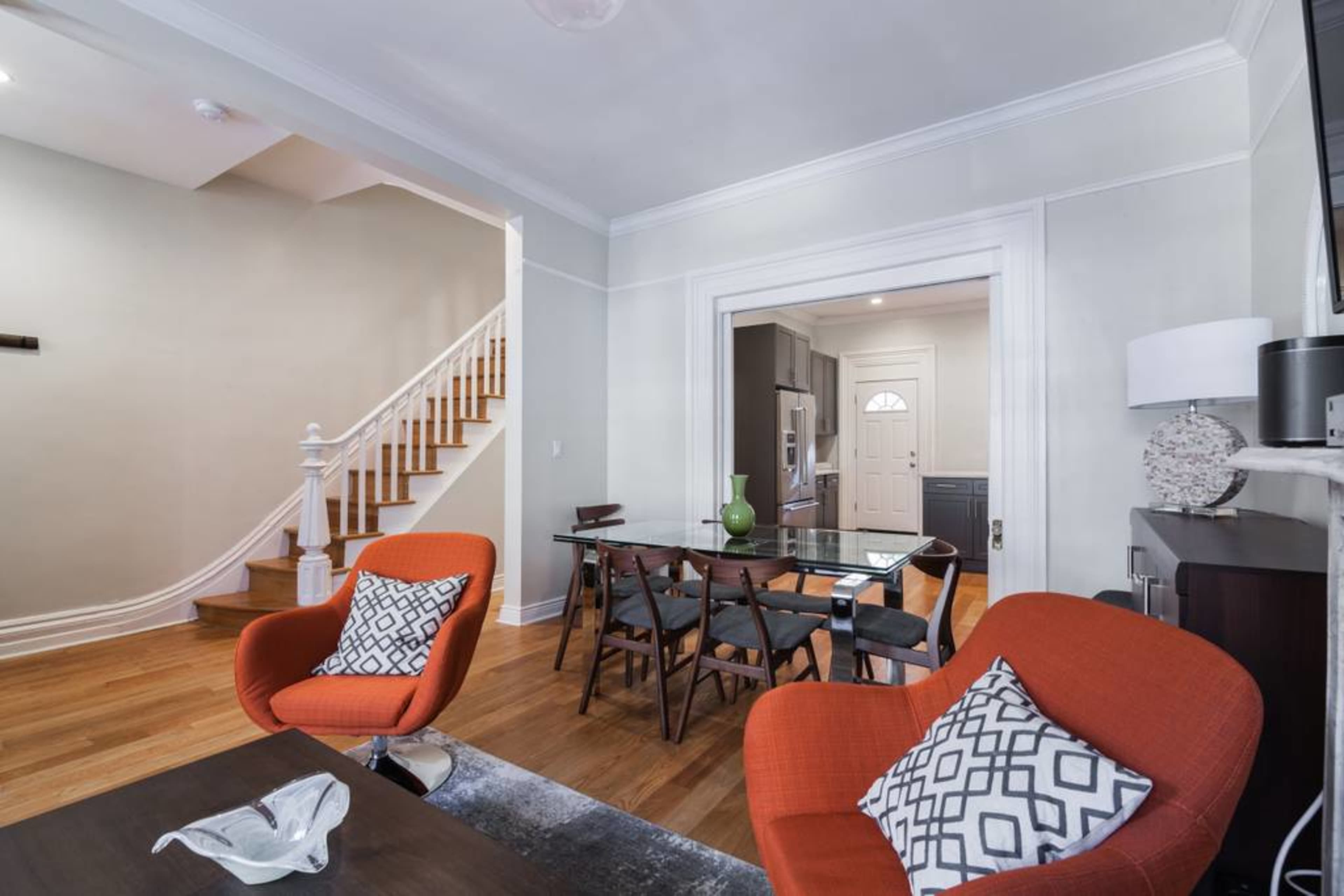 A living area with two orange chairs, a glass dining table, and a staircase leading to a second floor.