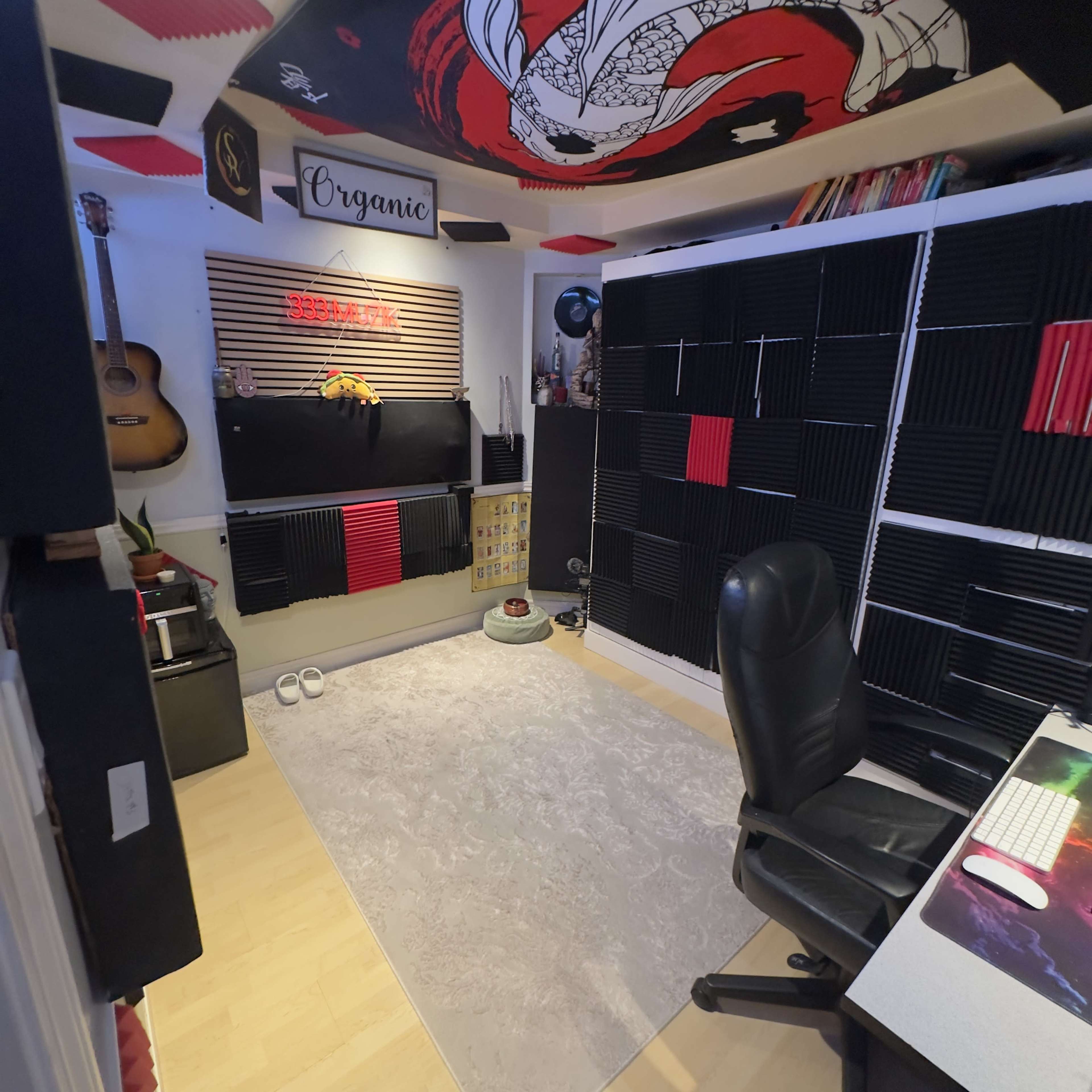 The image depicts a small recording or music studio with soundproofing panels on the walls, a desk with a computer, and a guitar hanging on the wall.