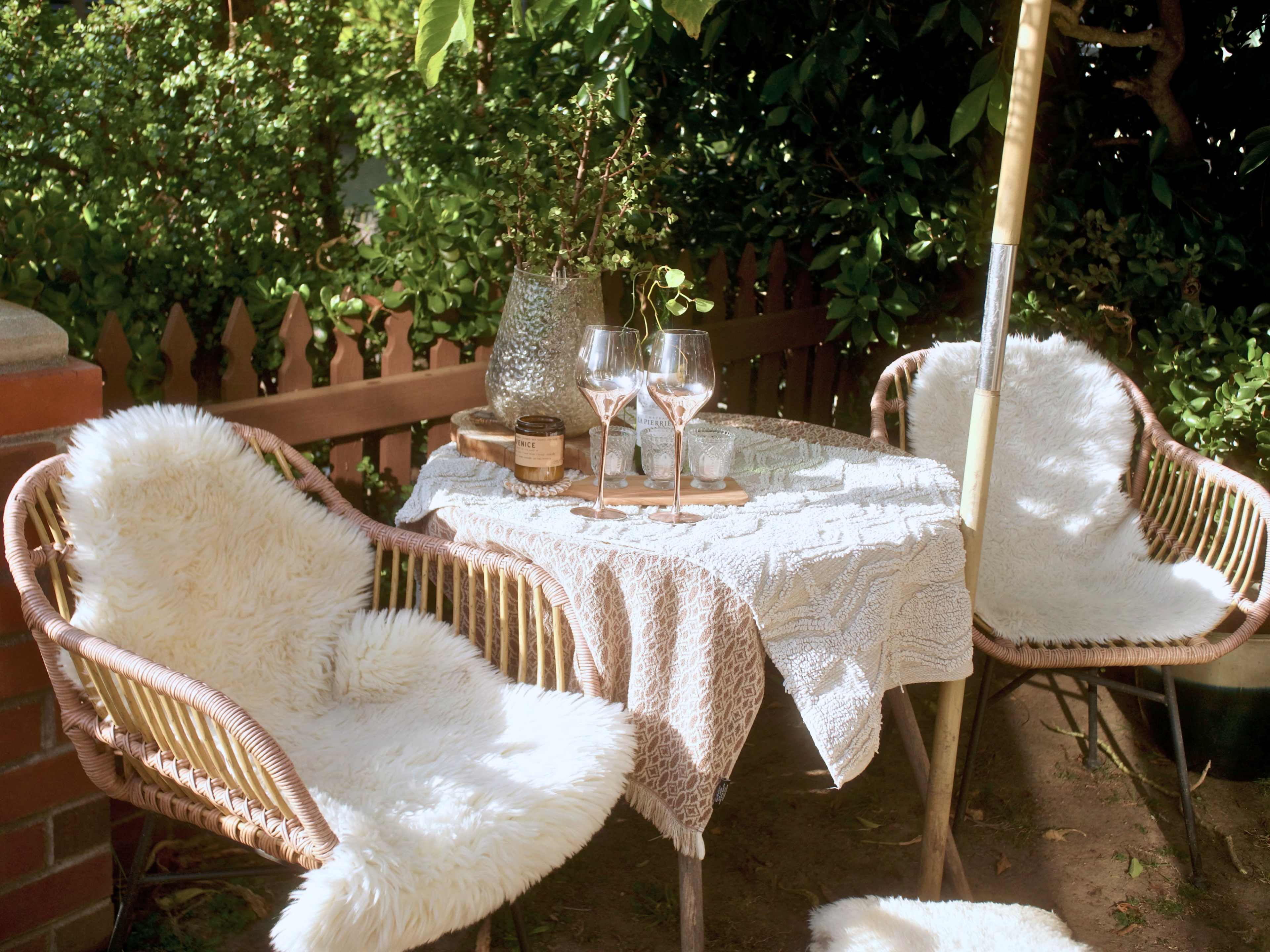 A small outdoor seating area features two wicker chairs with fur cushions, a table set with two glasses of rosé wine, and decorative items under a leafy canopy.