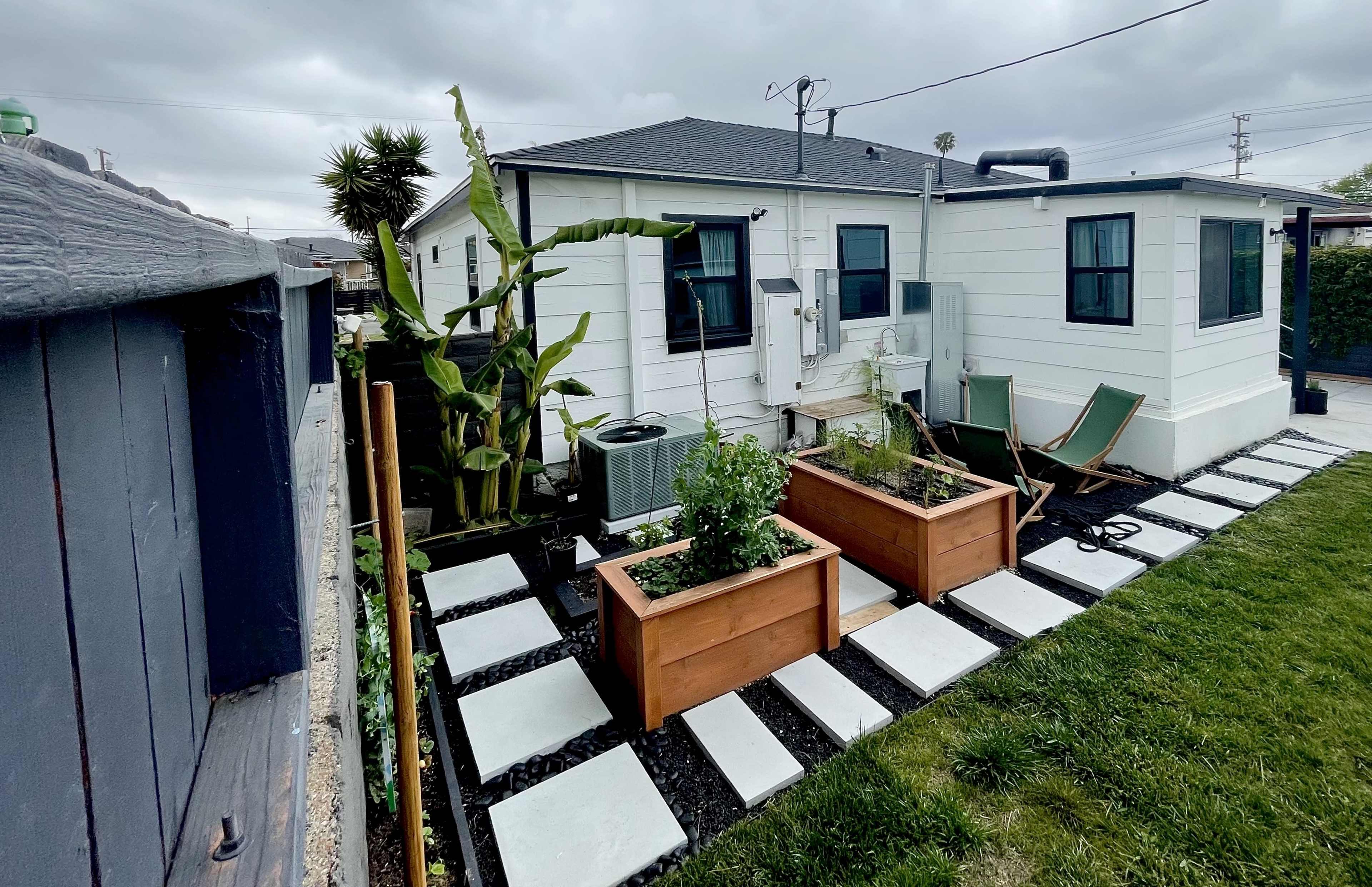 The image shows a backyard with two raised garden beds, stone pavers, and a small seating area beside a house.