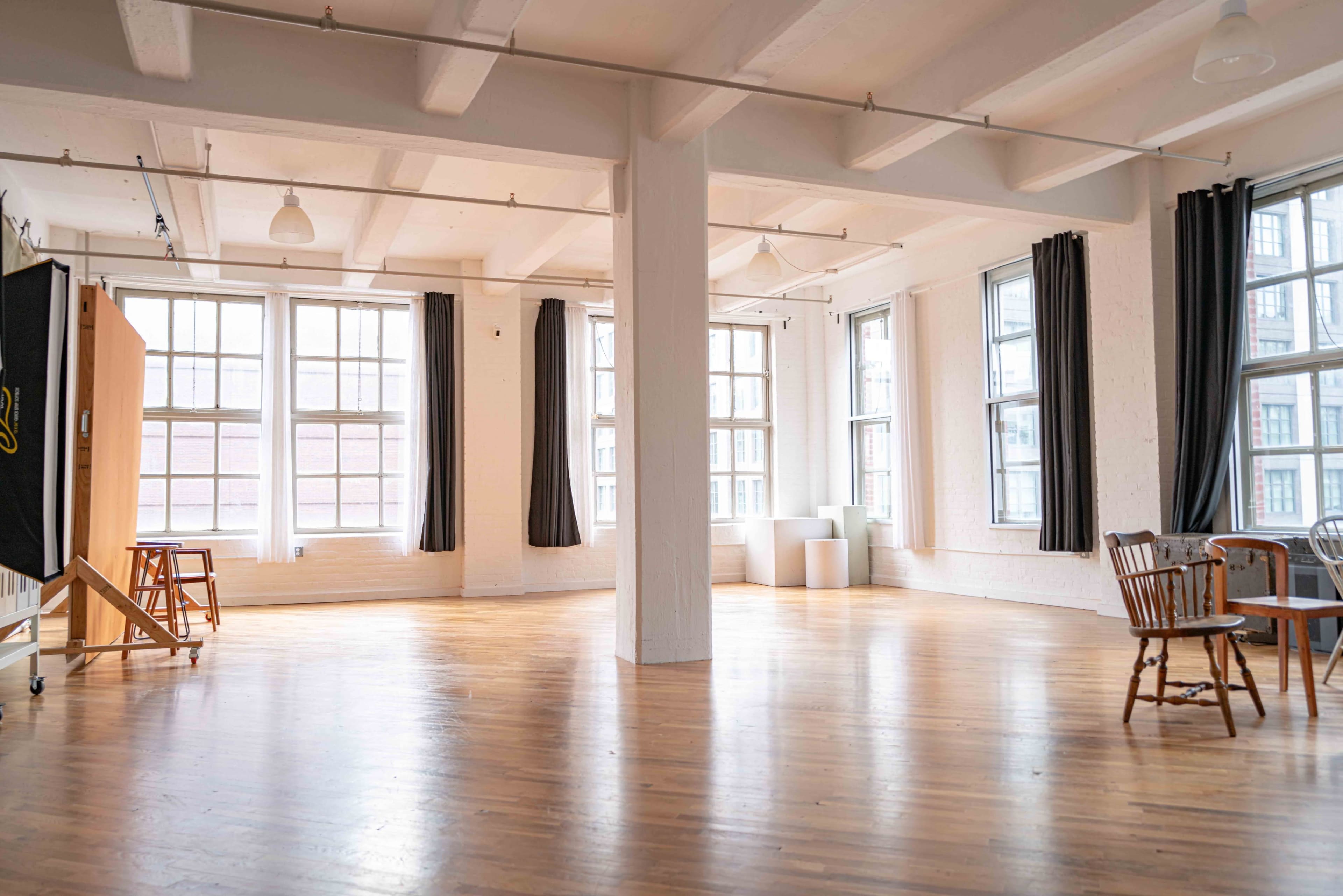 A spacious, well-lit room features large windows and wooden flooring, with a table and chairs near the corner.