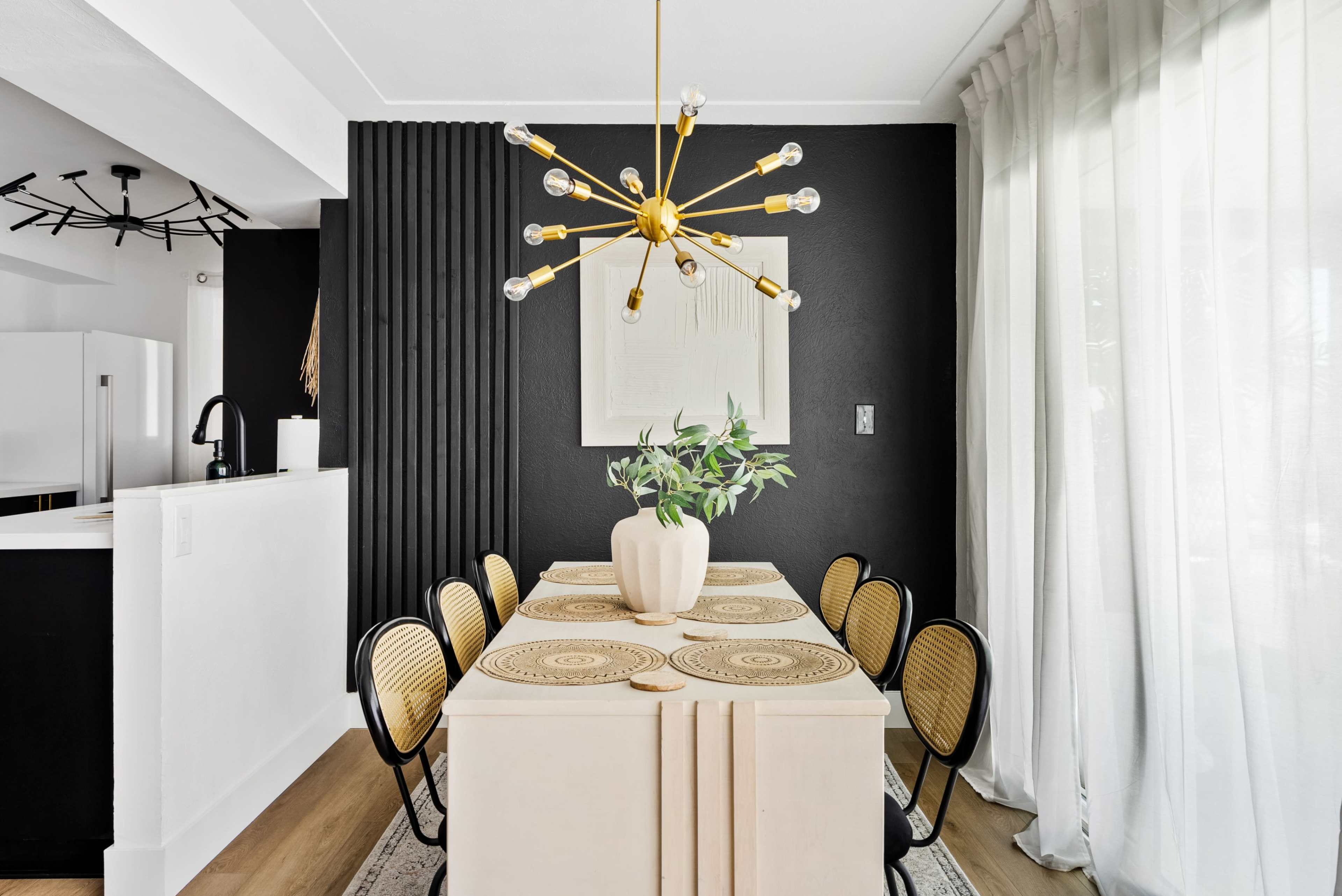 The image shows a modern dining area with a long table, eight black chairs, a gold starburst chandelier, and a large potted plant on the table, all set against contrasting black and white walls.