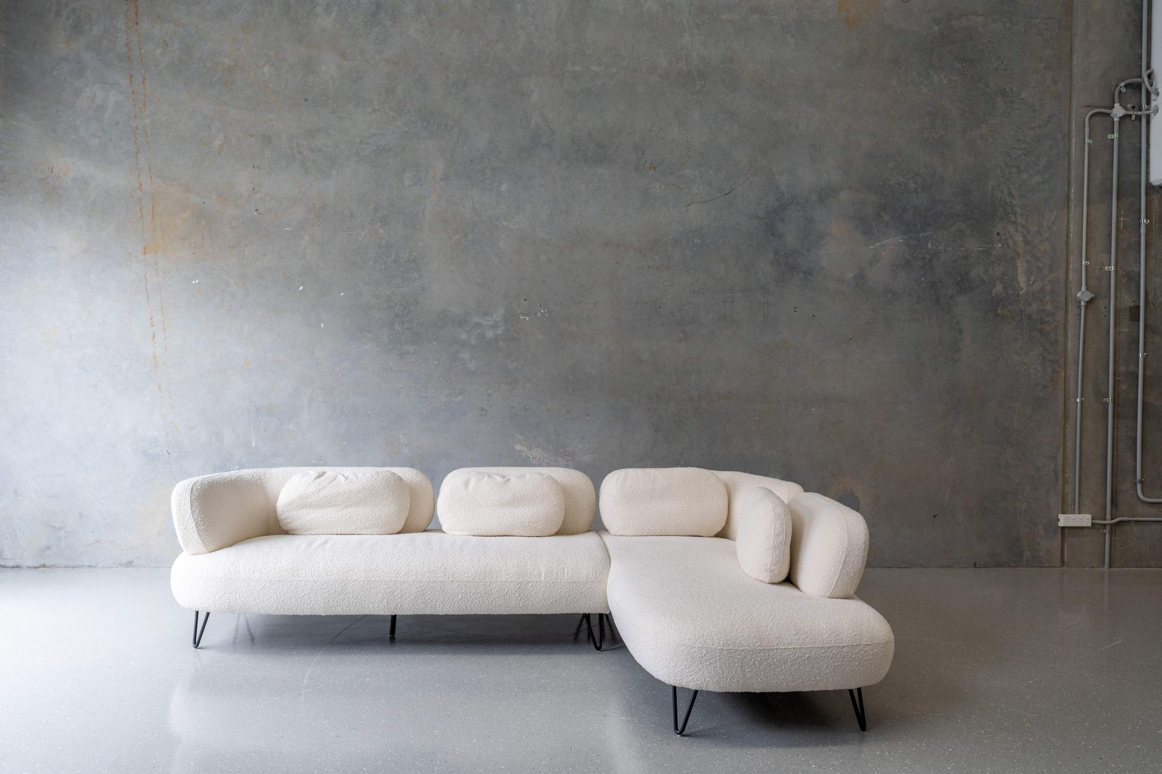 A light-colored, curved sectional sofa with rounded cushions is positioned against a textured gray wall.