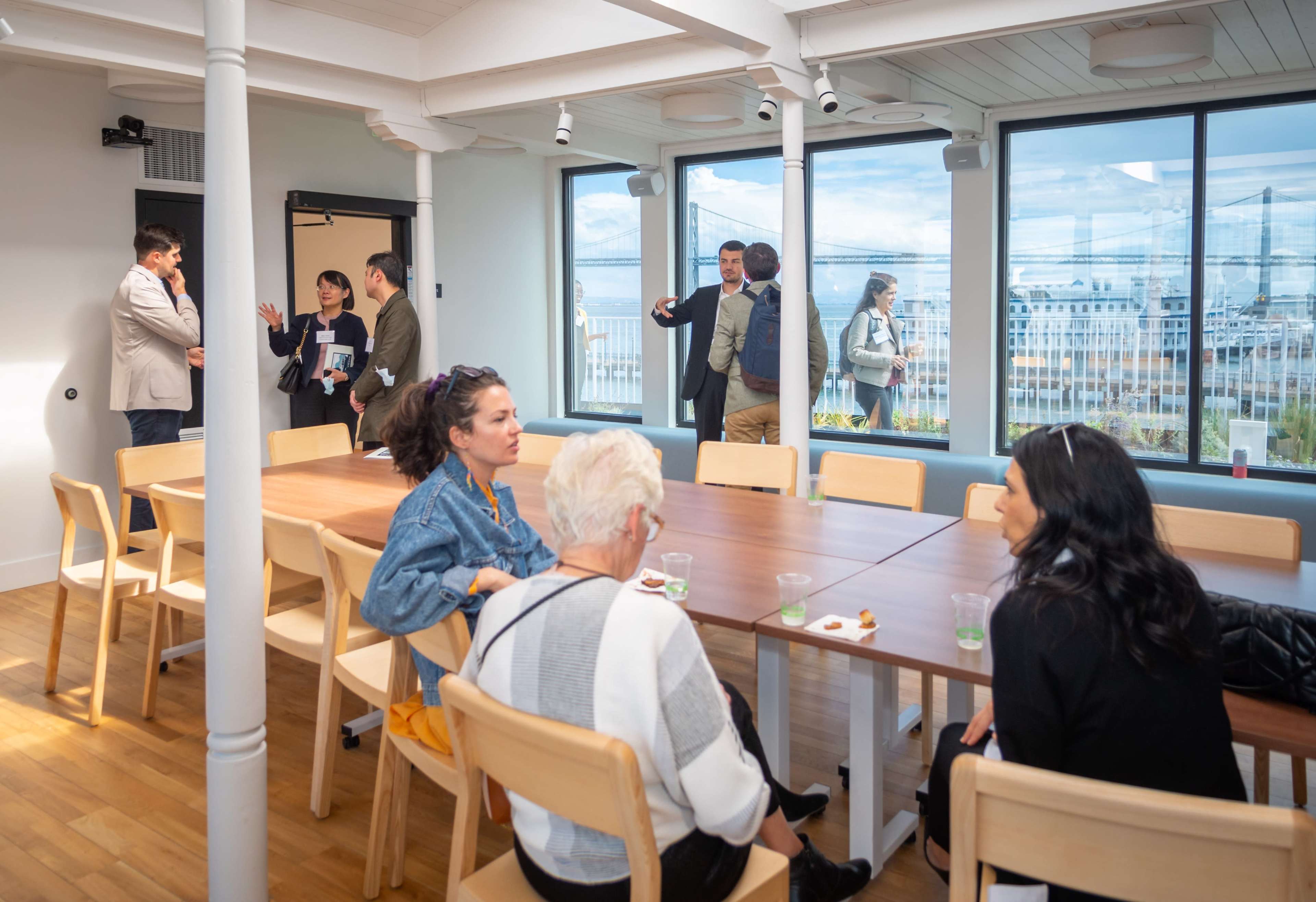 A group of people converses around a long table in a well-lit room with large windows overlooking a cityscape.