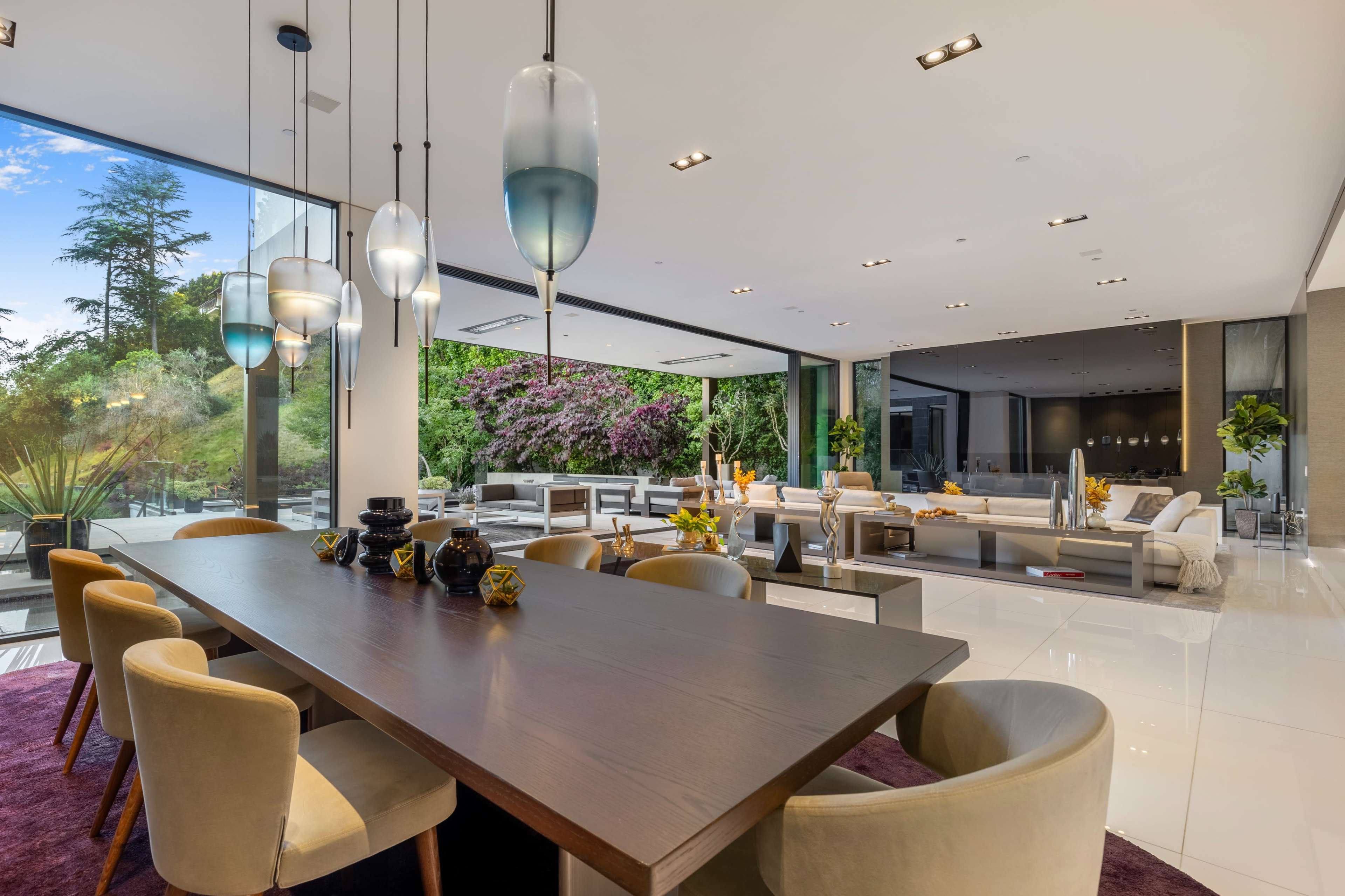 A modern dining area features a large wooden table surrounded by upholstered chairs, with large glass windows showcasing a garden view.