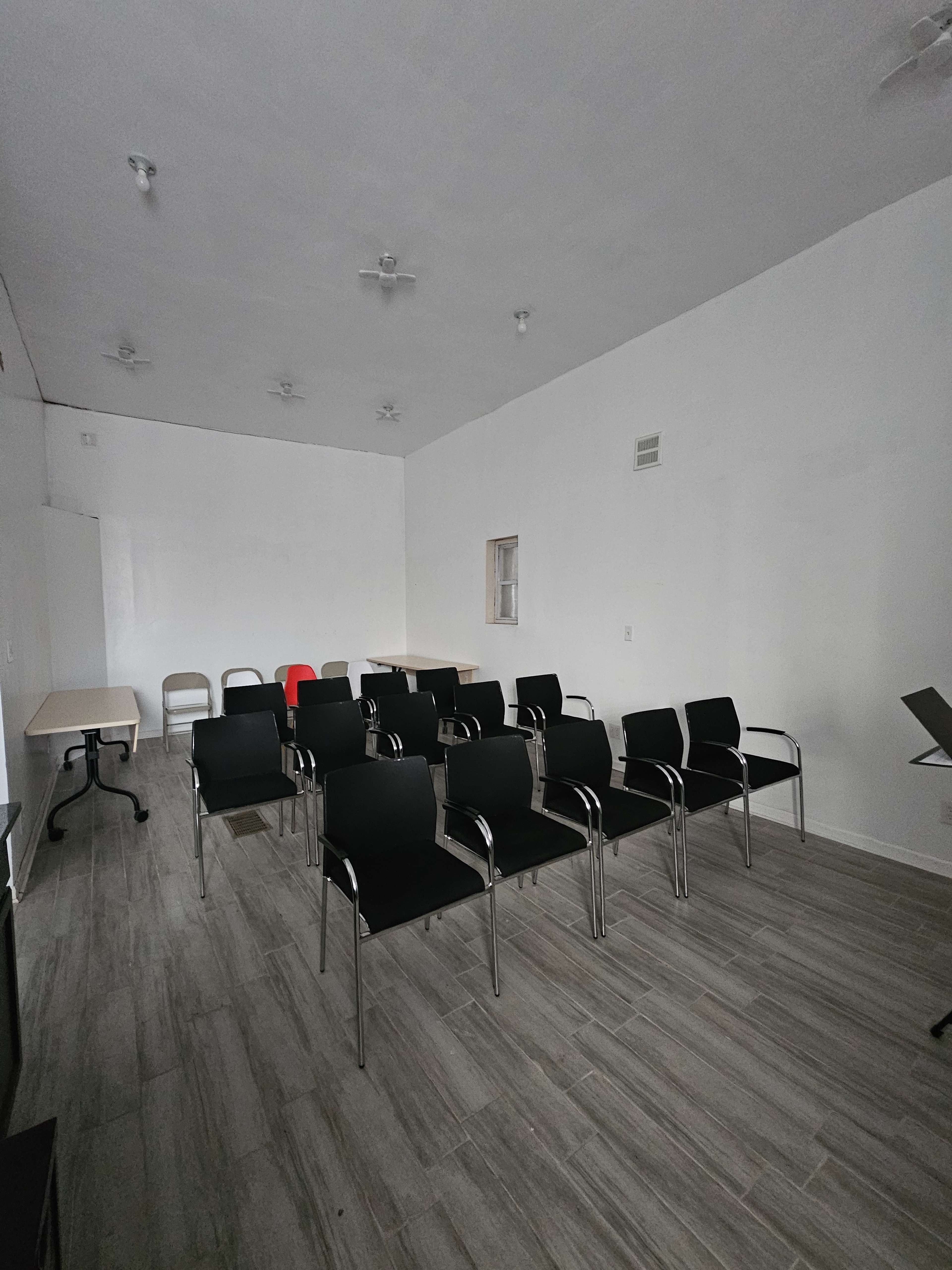 The image shows an empty meeting room with several black chairs arranged in rows and a few tables along the walls.